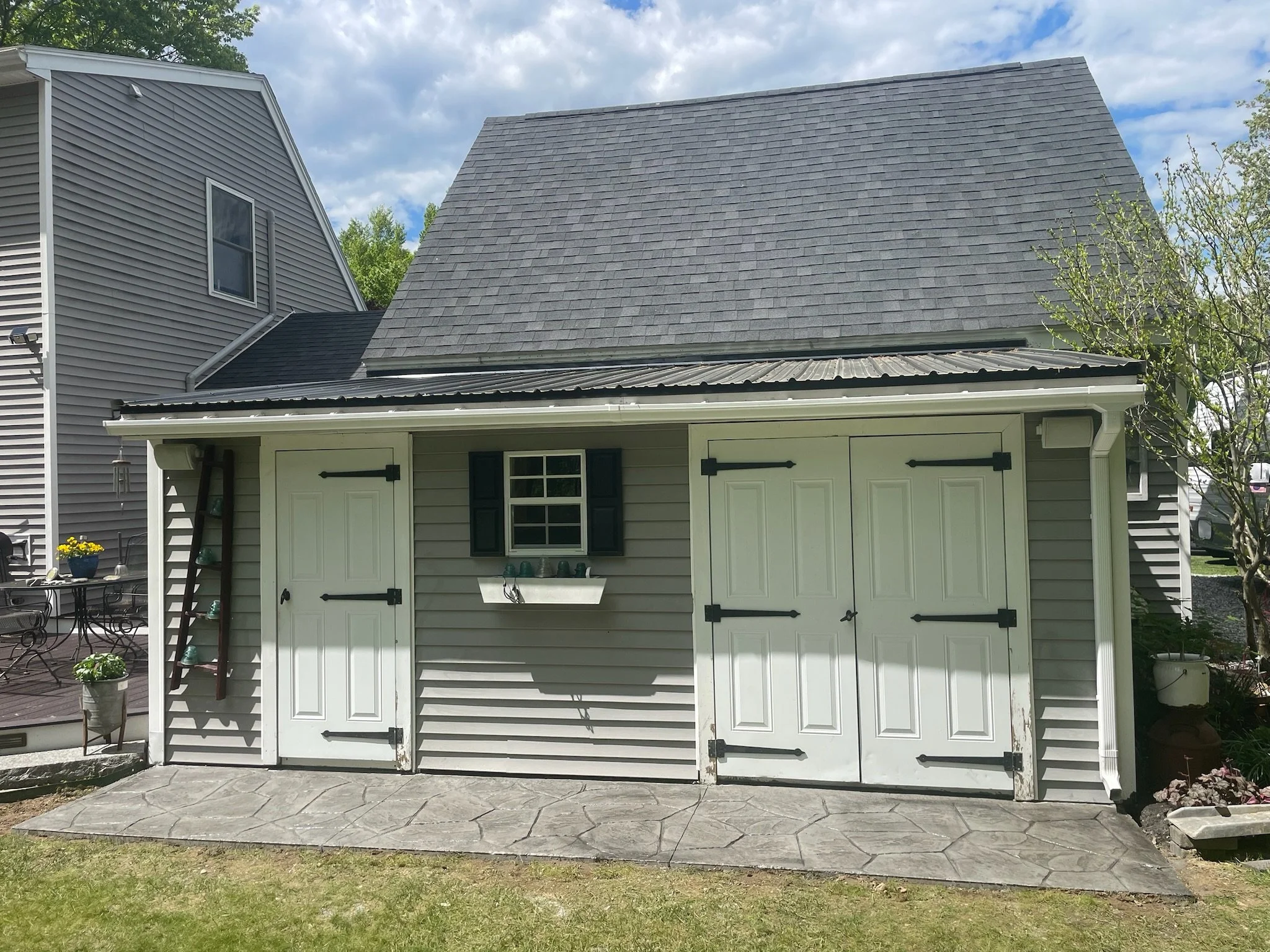 Backyard shed with gray siding, white double doors, a window with black shutters, and decorative black hardware, on a stamped concrete stone patio with a green lawn in the foreground in Meredith New Hampshire.