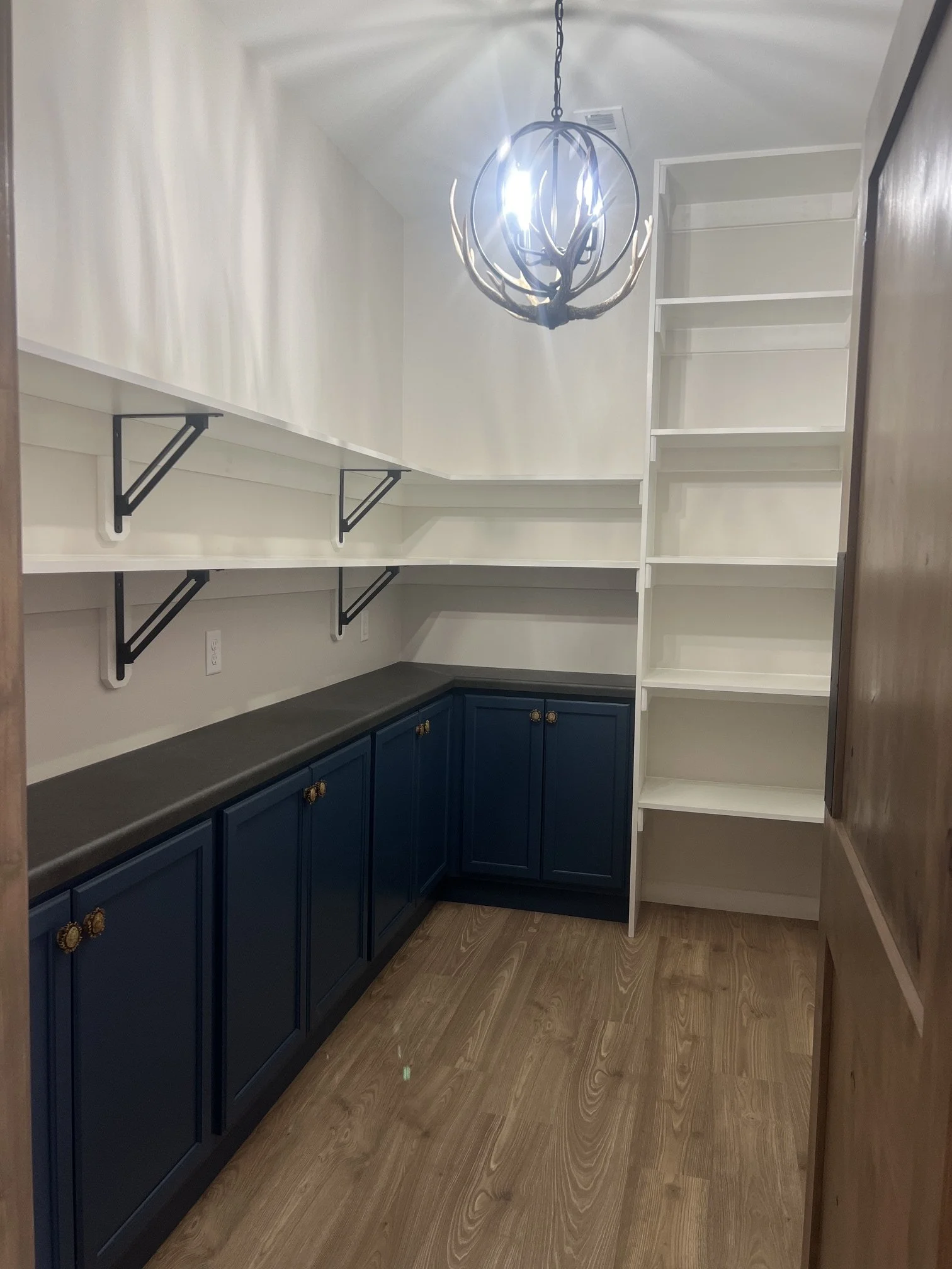 Custom walk-in pantry with navy cabinetry, white open shelving, wood flooring and antler-style light fixture.