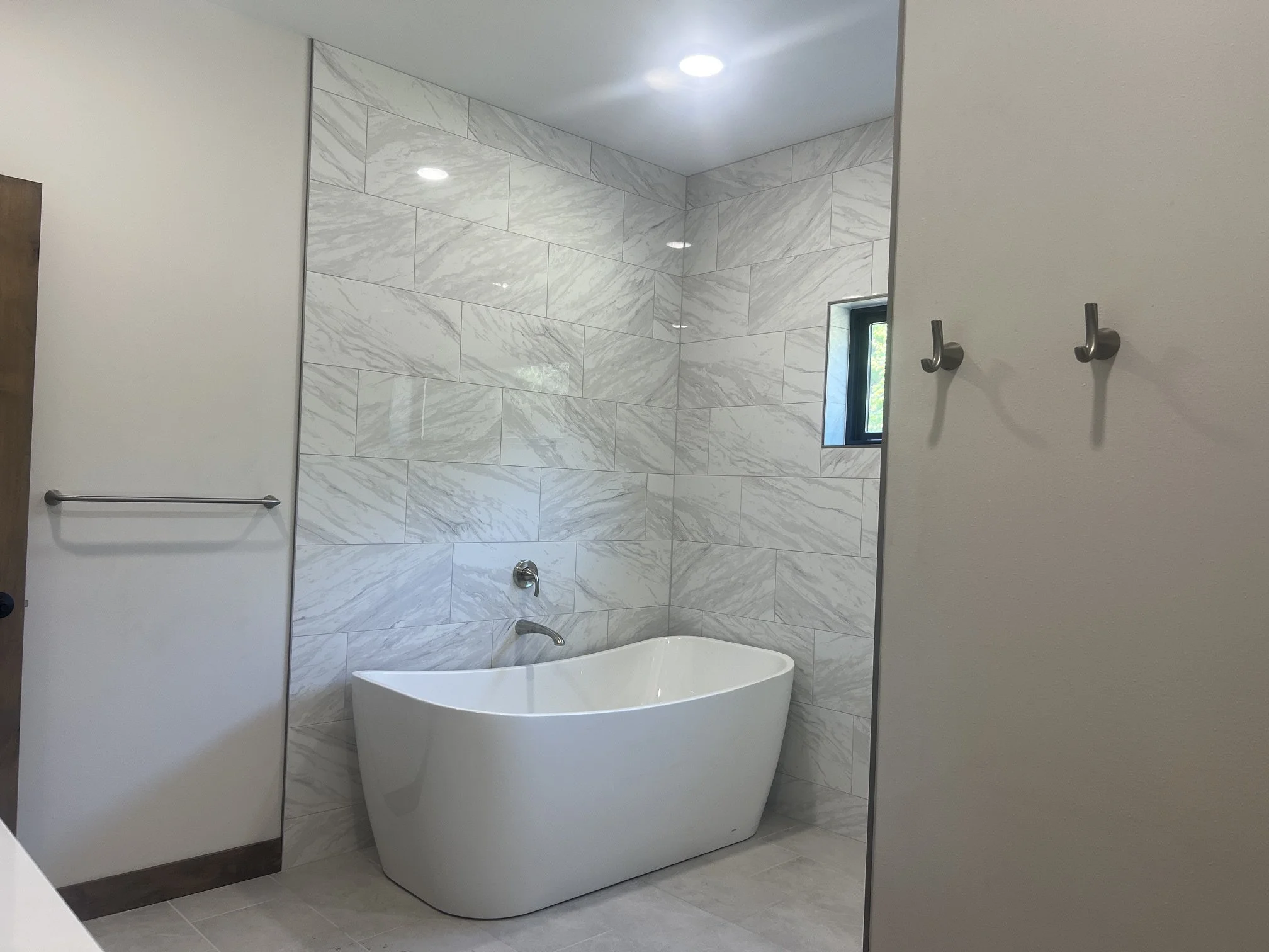 Freestanding soaking tub surrounded by white marble-look tile in a bright custom bathroom.