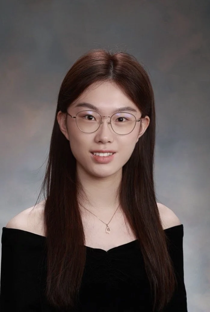 Portrait of a young woman with long brown hair, glasses, and a black off-the-shoulder top, against a grey background.