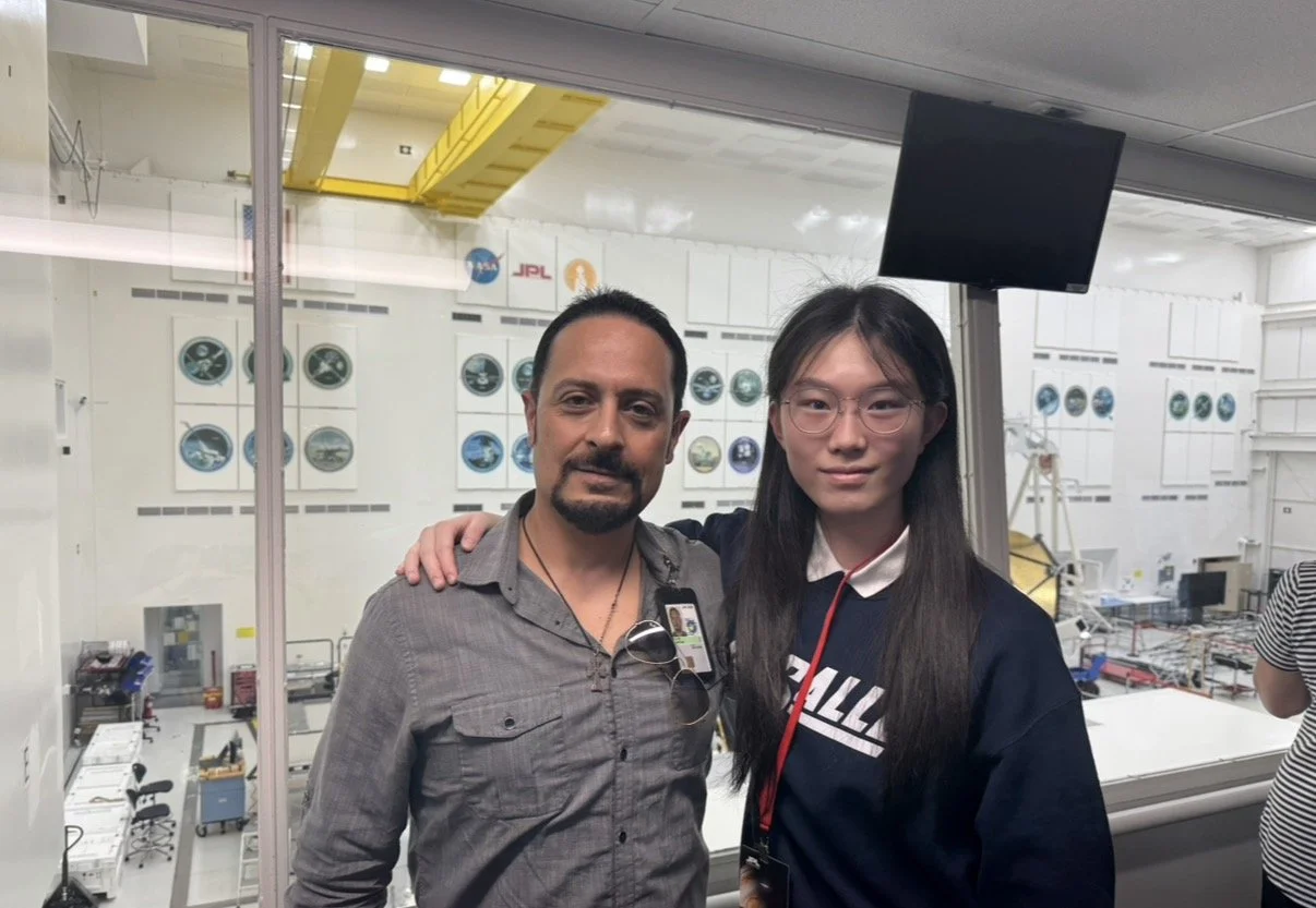 Two people, a man and a woman, standing together in front of a large glass window overlooking a NASA space lab or cleanroom with various equipment and wall displays, posing for a photo.