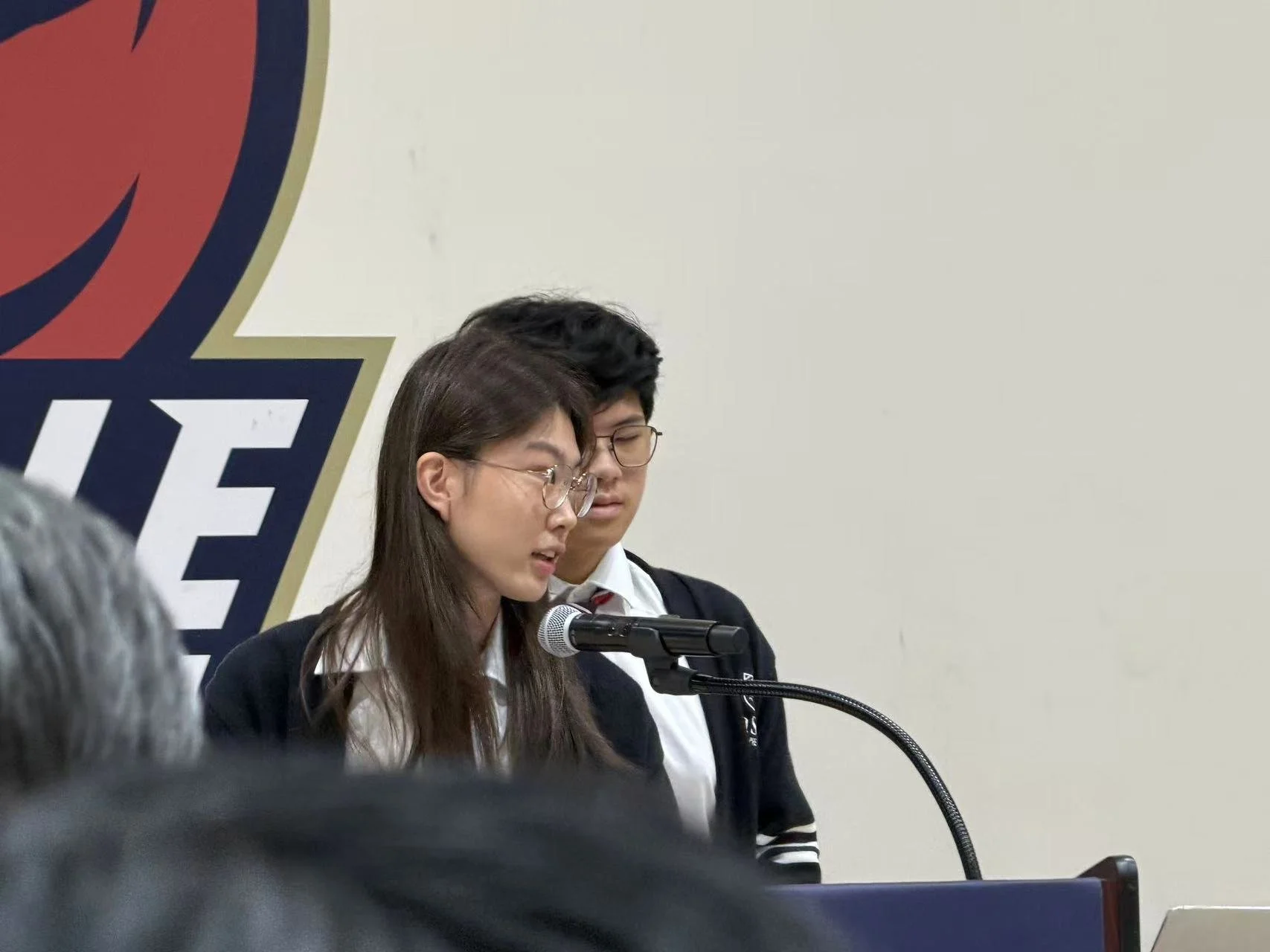 Two students, a girl and a boy, standing at a podium, speaking into a microphone in a room with a poster or banner featuring a logo behind them.