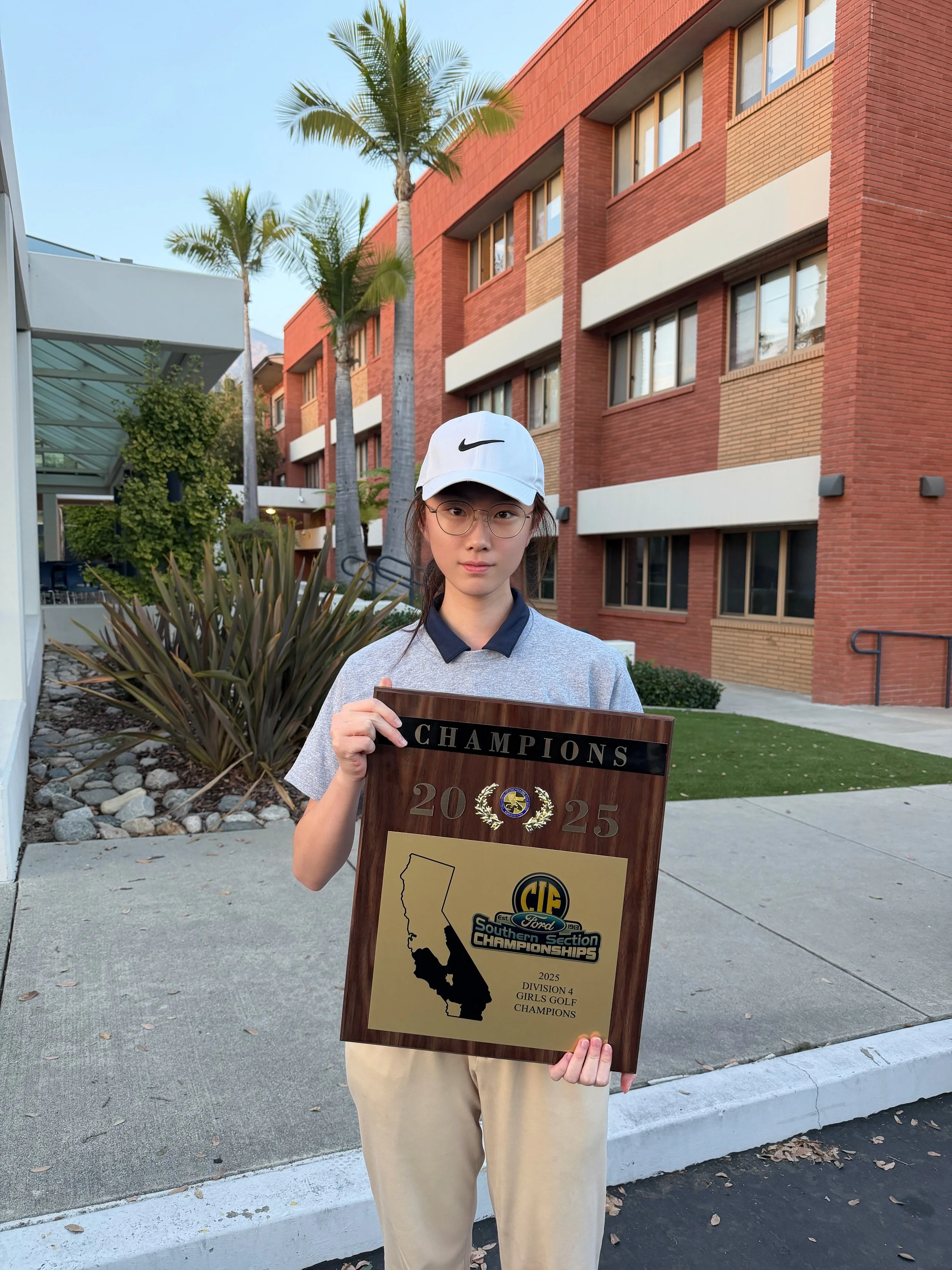 A young woman wearing glasses and a white Nike cap, standing outdoors in front of a modern brick building with palm trees, holding a plaque that reads 'Champions 2025, CIF Southern Section Championships, Division 4 Girls Golf Champions'.