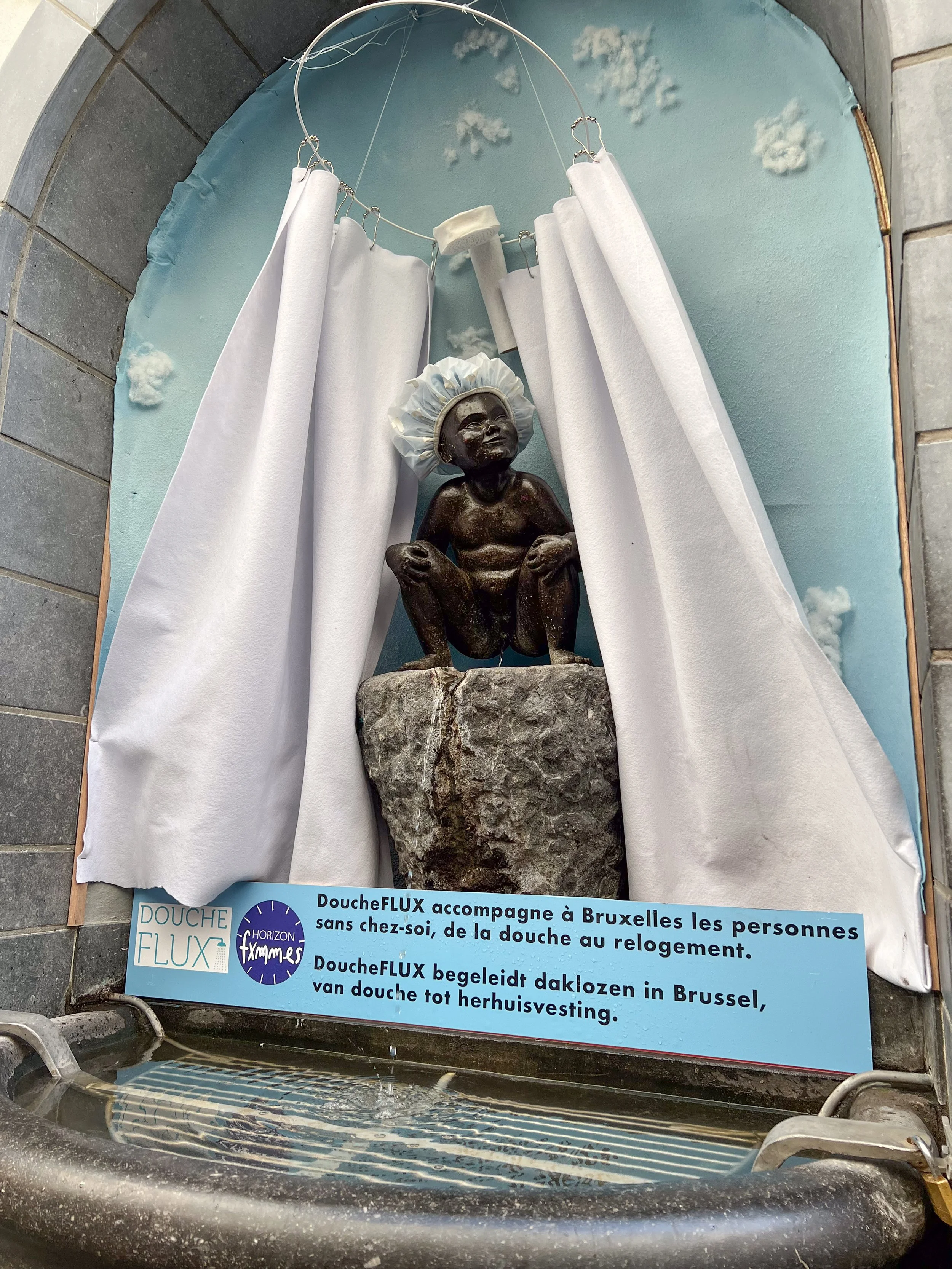 The current decoration for "Jeanneke Pis" is by DoucheFlux, an organization working to end homelessness. The caption translates to "In Brussels, our support services ensure that people feel at home—from the shower to their housing."