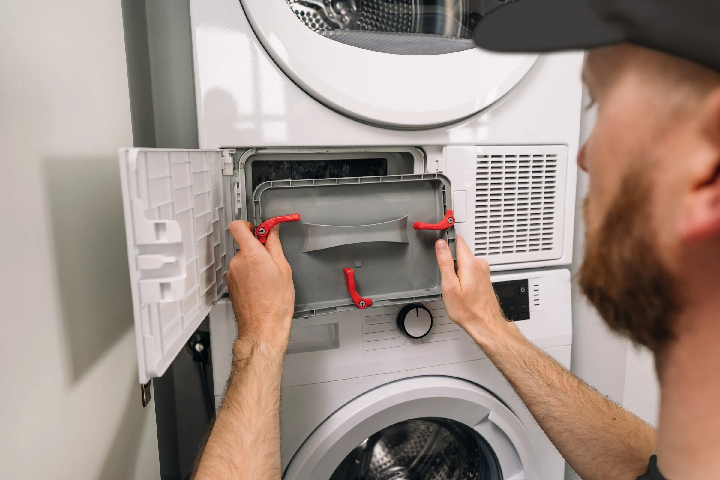 A man with a beard and hat opening the lint filter of a front-loading washing machine.