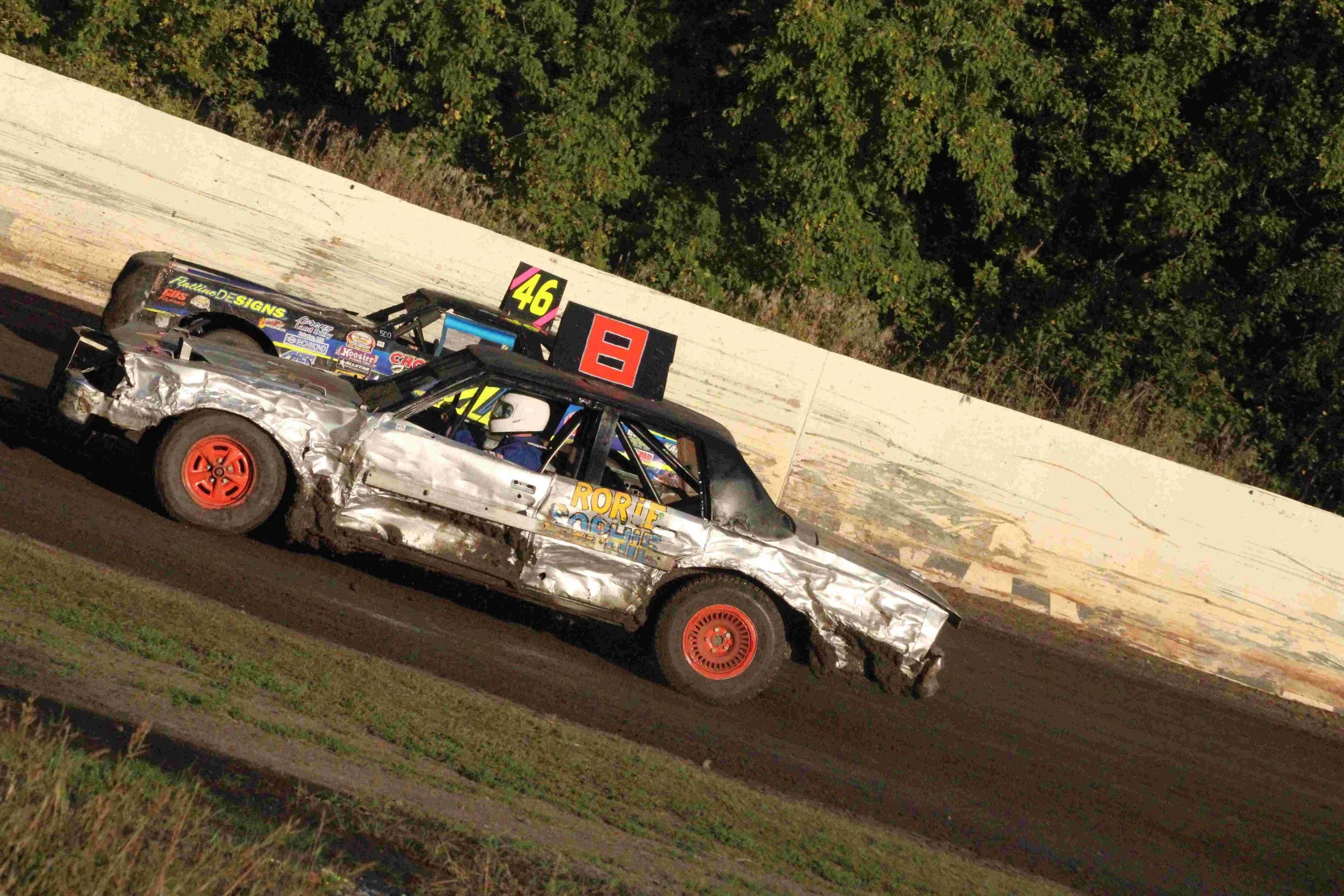A damaged race car with a driver wearing a helmet on a dirt track, alongside another race car, with a white concrete barrier and green trees in the background.