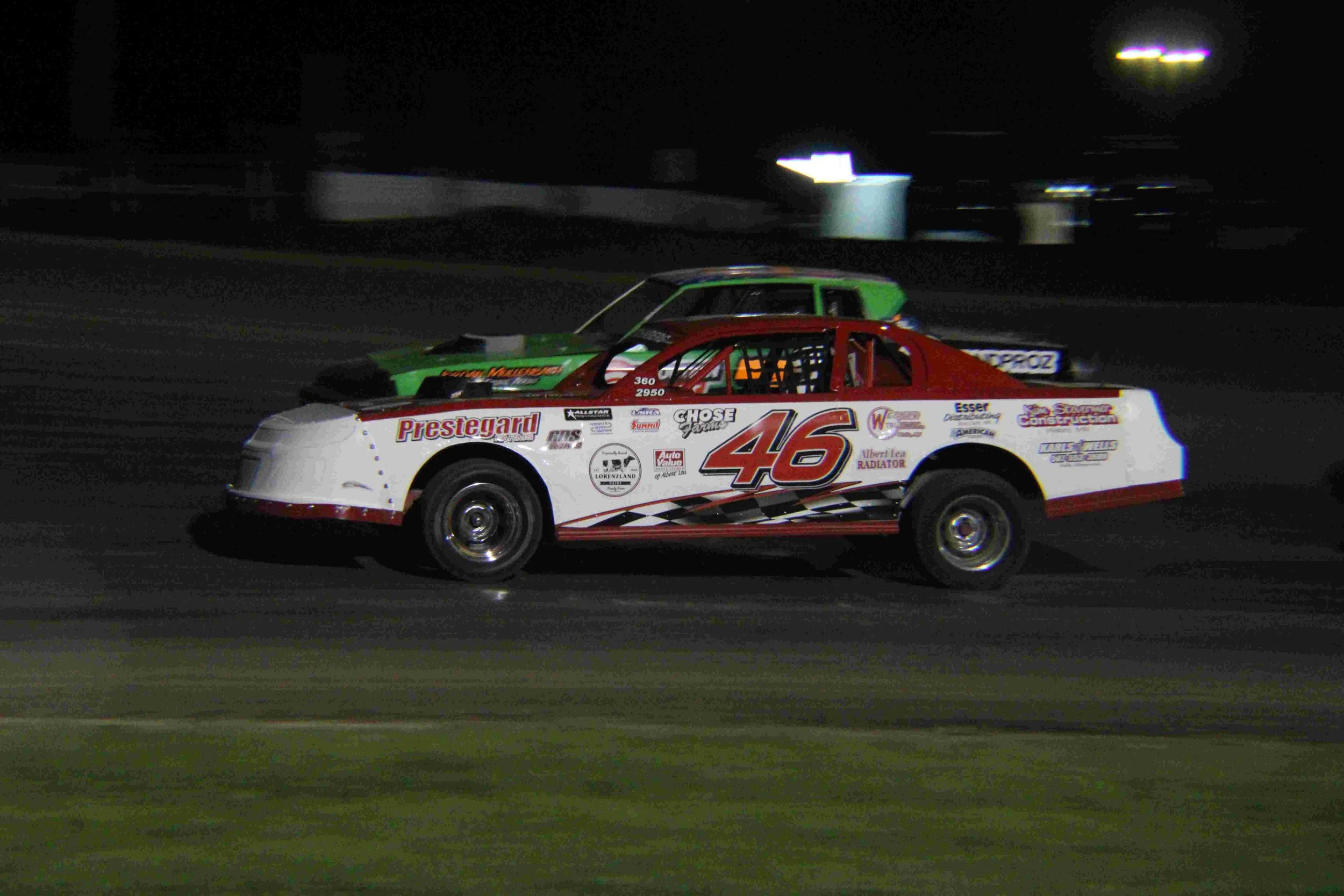 Nighttime dirt track racing with two race cars; white car with red and black accents, number 46, and green car in the background.
