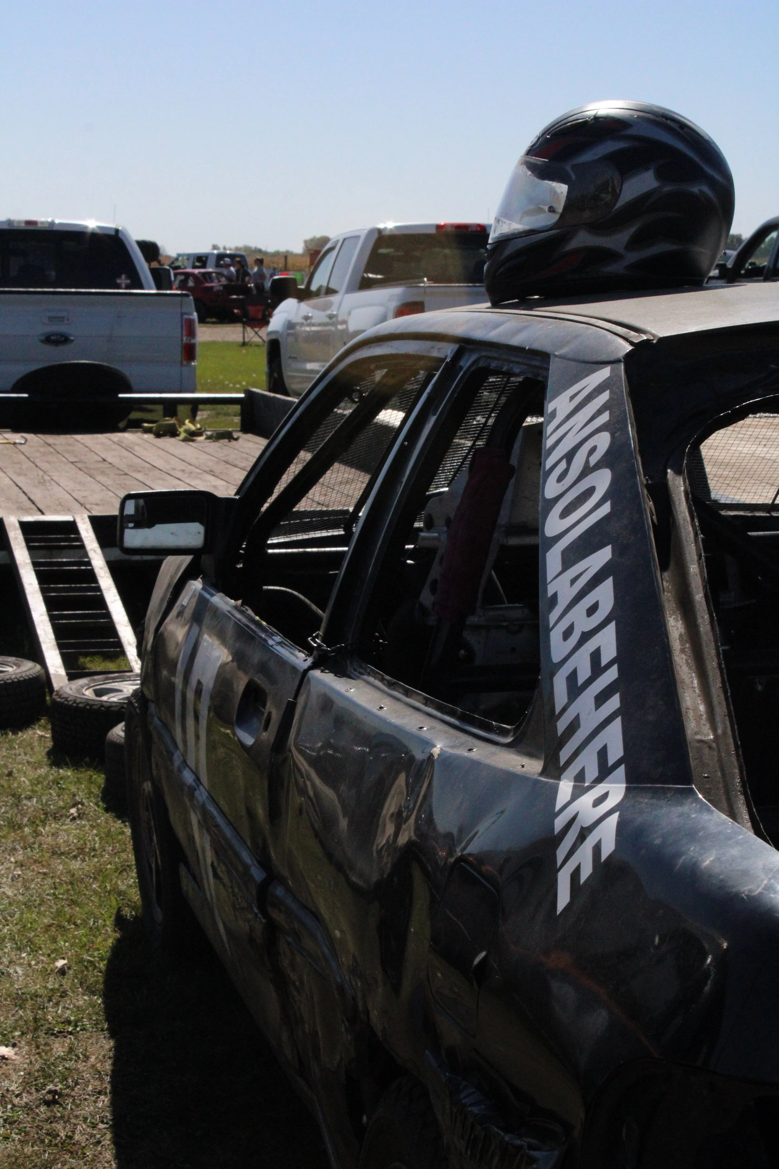 Wrecked race car with a helmet on top, parked on a grassy area, with tents and vehicles in the background.