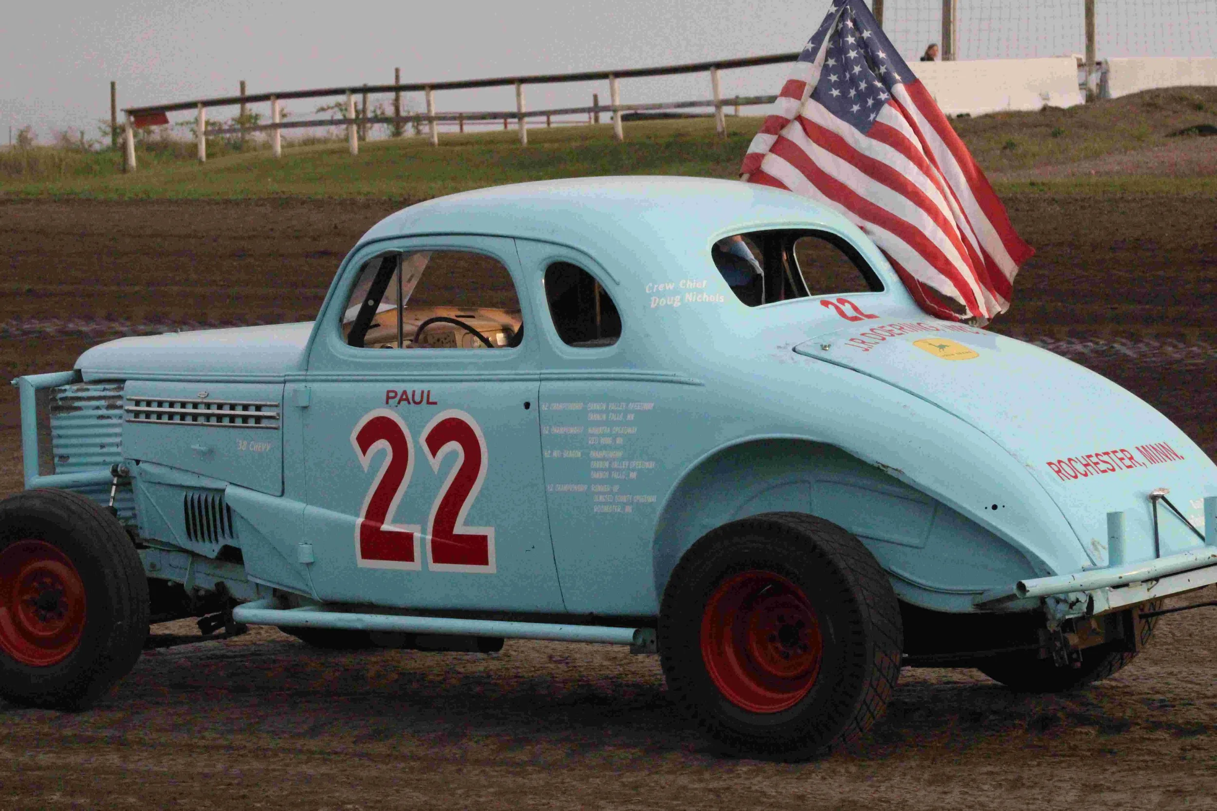 A vintage light blue racing stock car with the number 22 in red on its side, featuring an American flag mounted on the roof, on a dirt race track at Lansing, Minnesota.