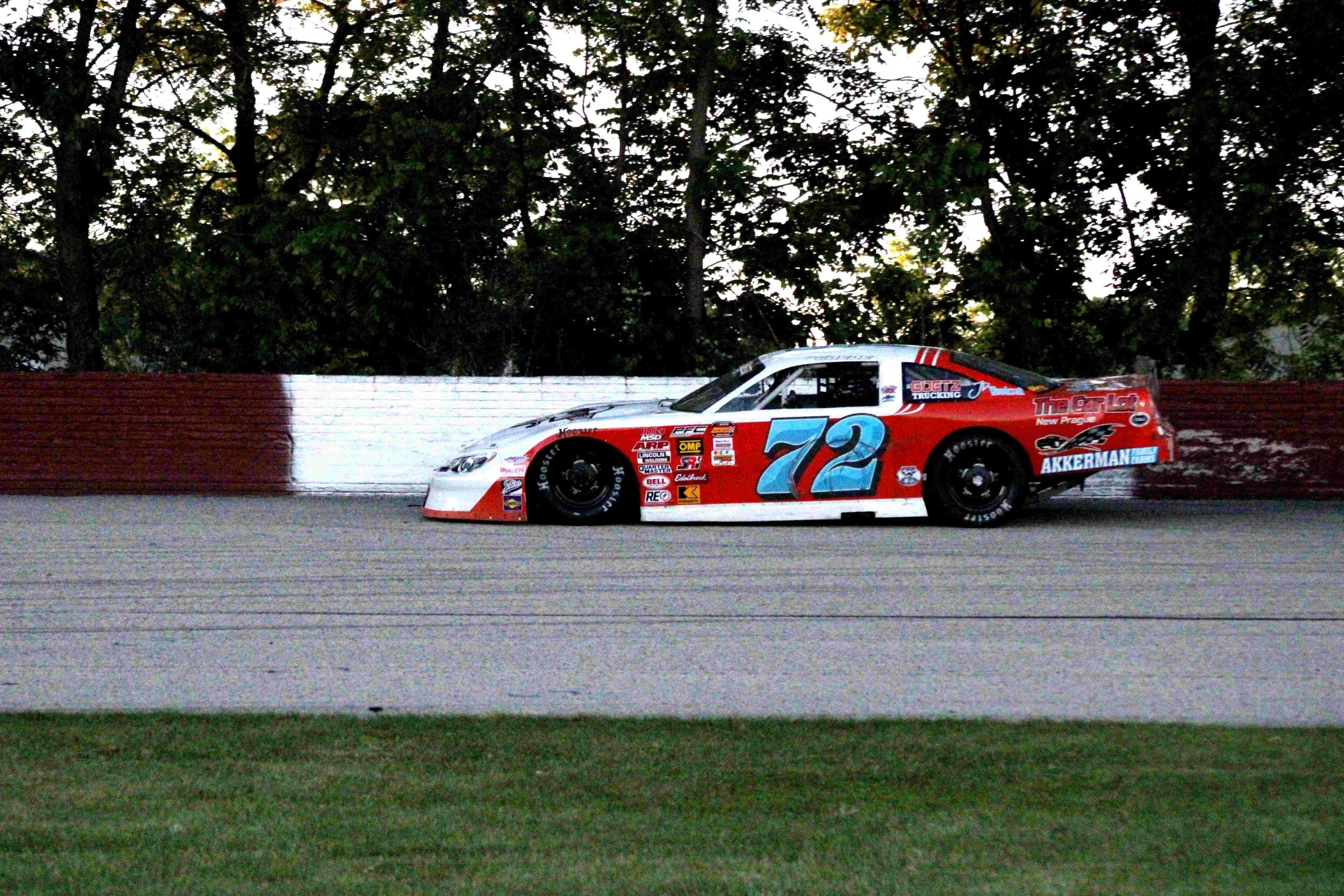 A red and white stock car with the number 72 on the side driving on a race track near a red and white barrier, with trees in the background.