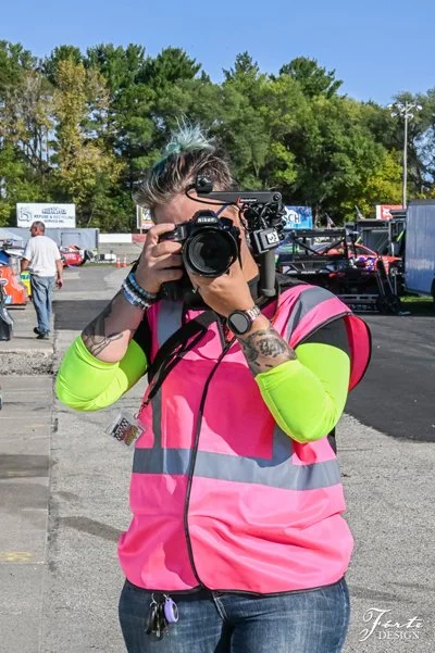 A woman with short hair and rainbow-colored highlights taking a photo with a camera, wearing a bright pink and yellow safety vest, standing outdoors in front of trees and parked cars.