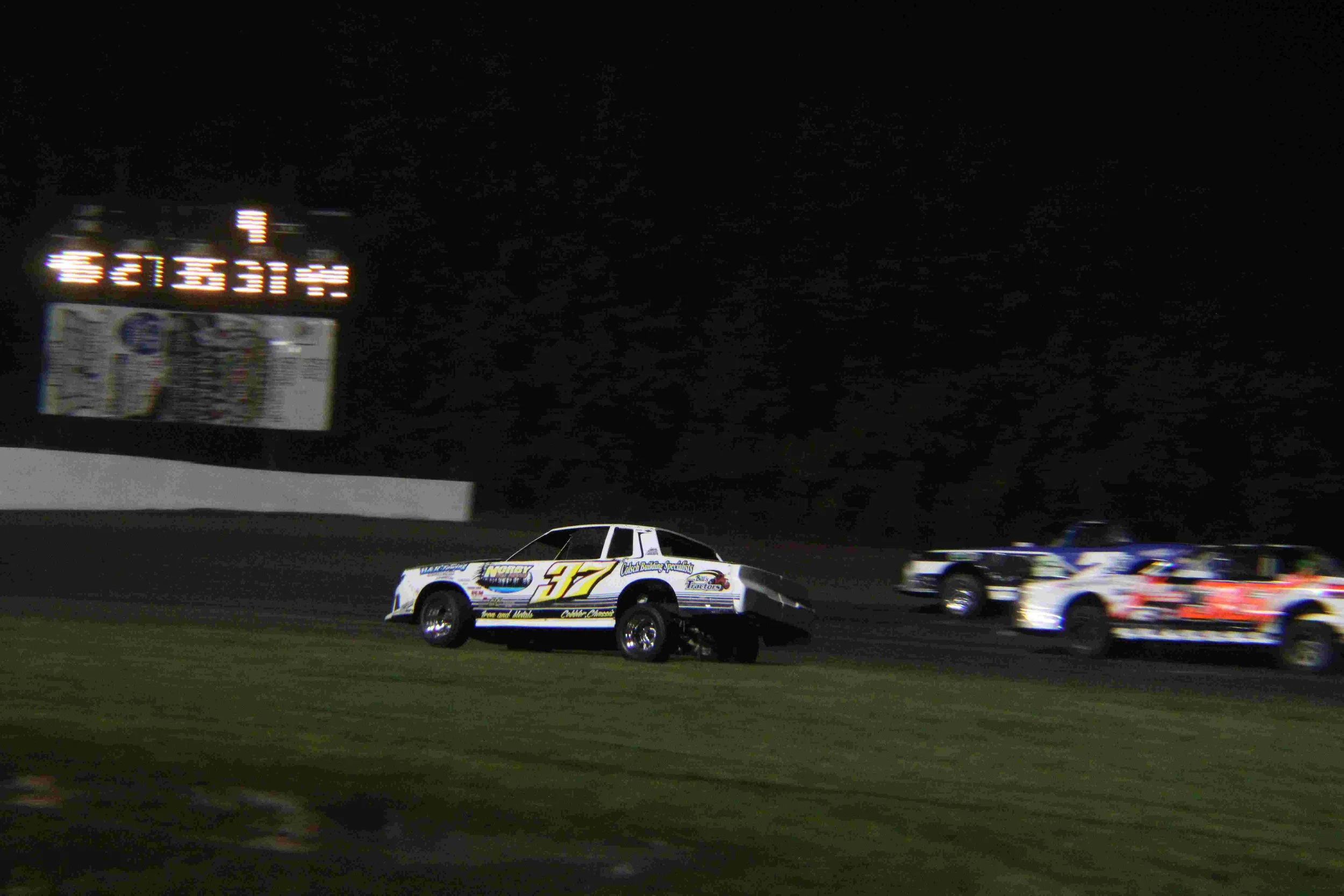 Nighttime dirt track race with three race cars, one white number 37, and a digital scoreboard displaying information.