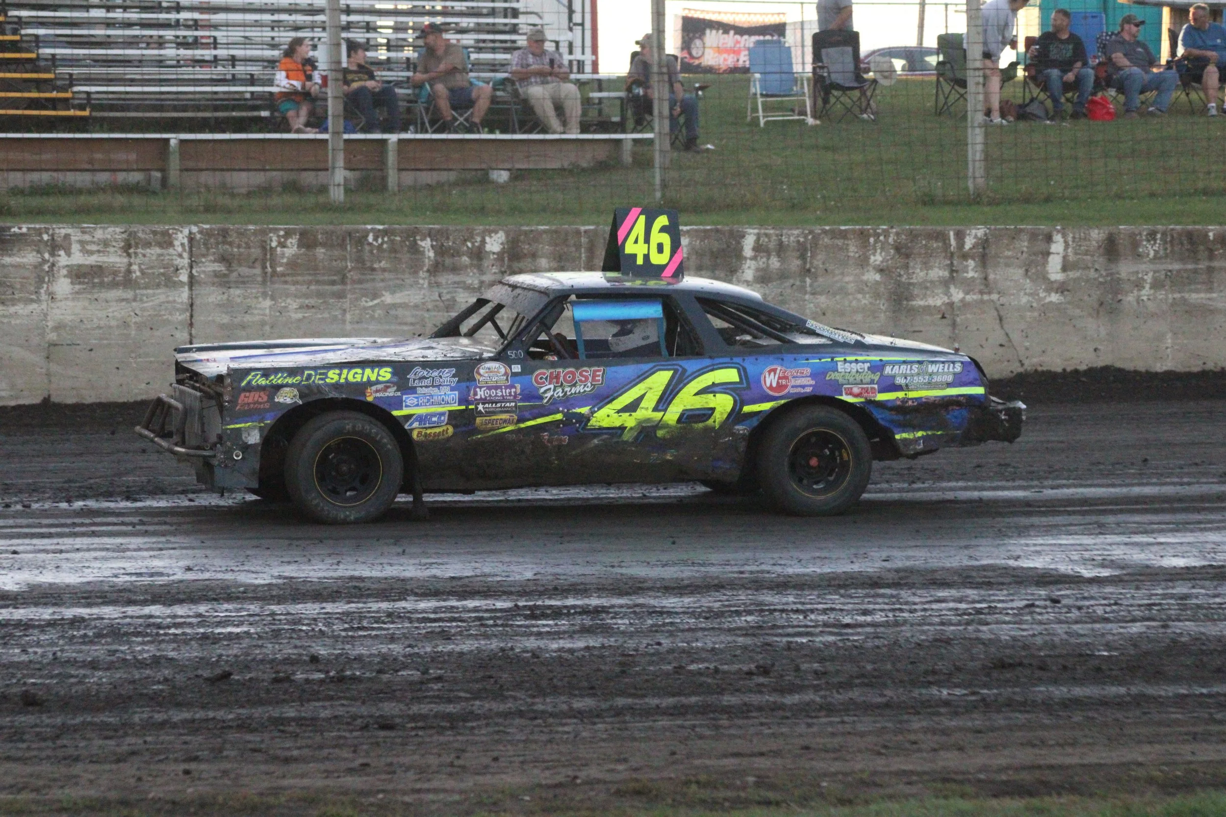 A blue race car with the number 46 on its side and a pink and green number 46 sign on top, driving on a dirt track with spectators in the background.