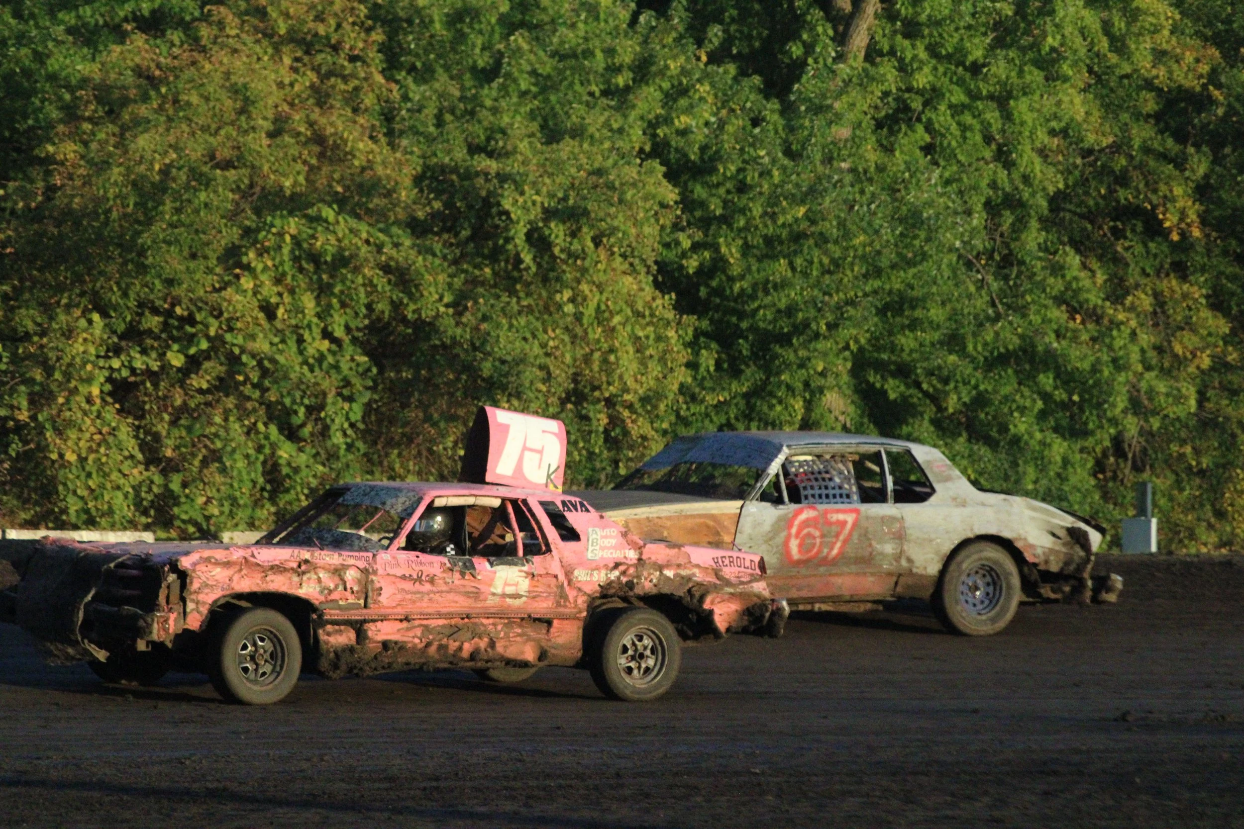 Two damaged racing cars on a dirt racetrack with green trees in the background, one pink and the other silver, with numbers 75 and 67.