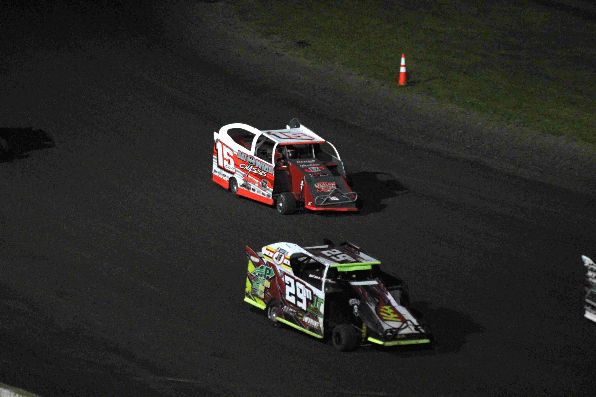 Two race cars on a dark dirt track, with an orange and white traffic cone in the background.