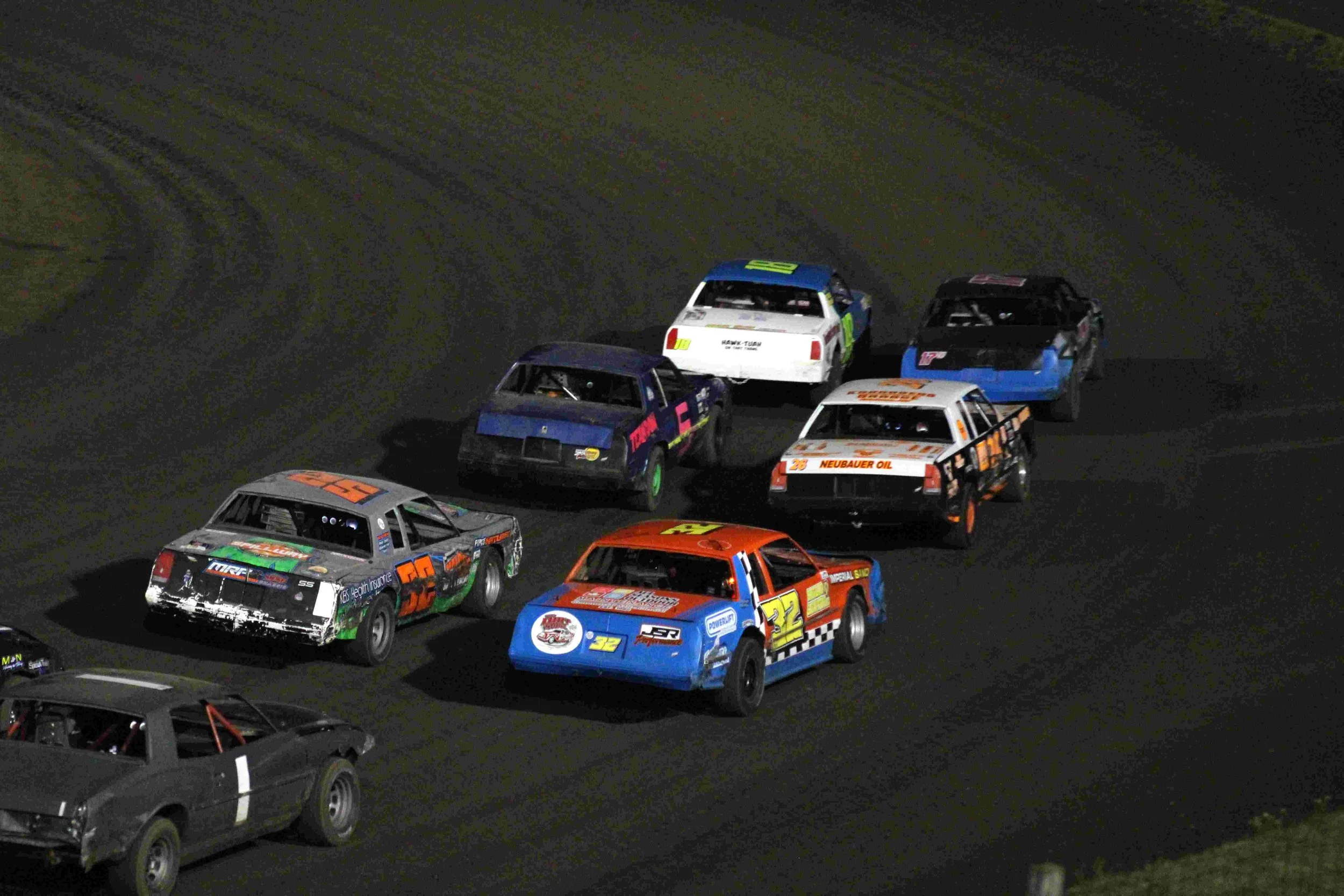 Group of stock cars racing on a dark asphalt track at night, making a turn with six cars in background, featuring various colors and sponsor decals.