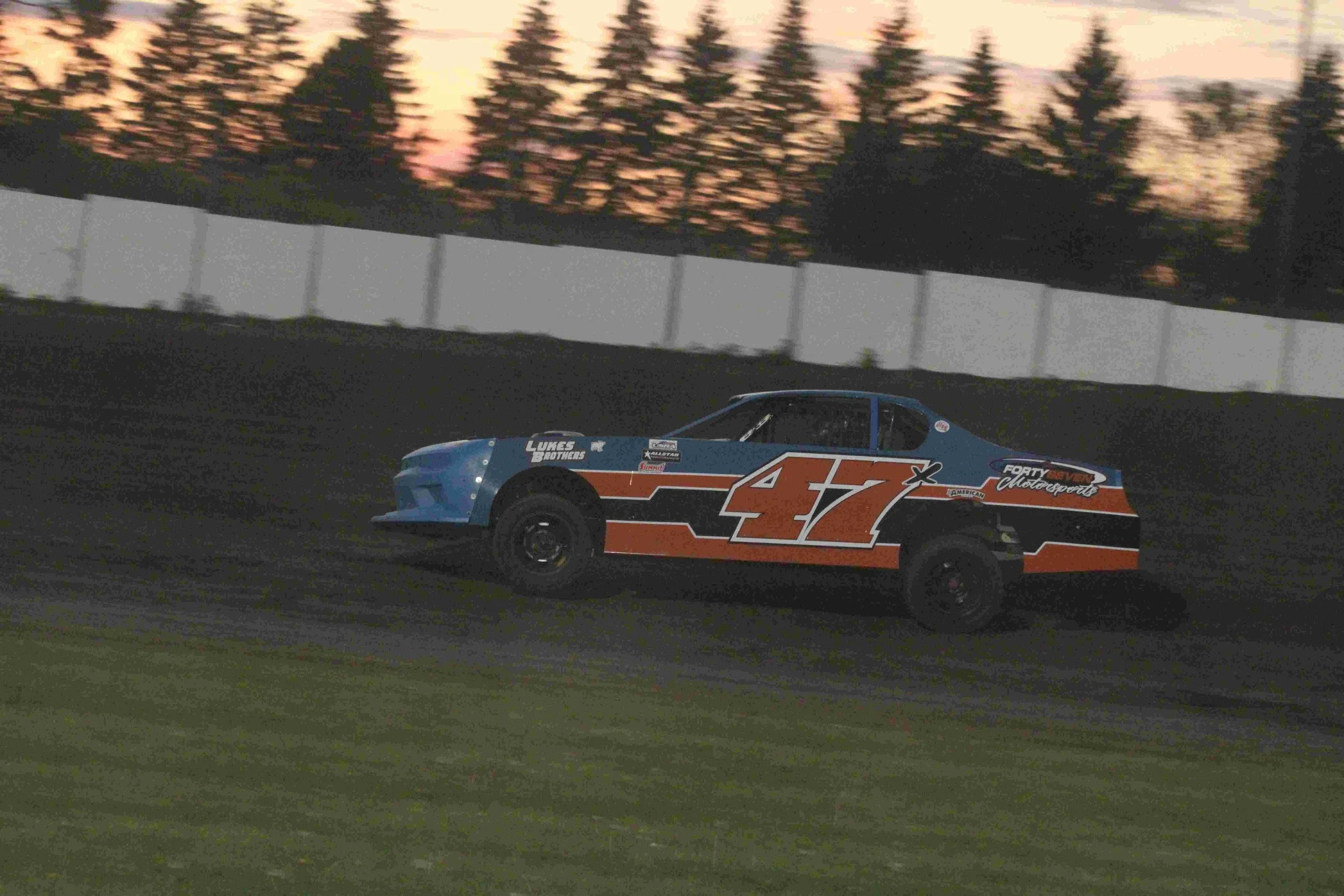 A race car with number 47 on the side speeding on a dirt track during sunset, with trees and a white fence in the background.