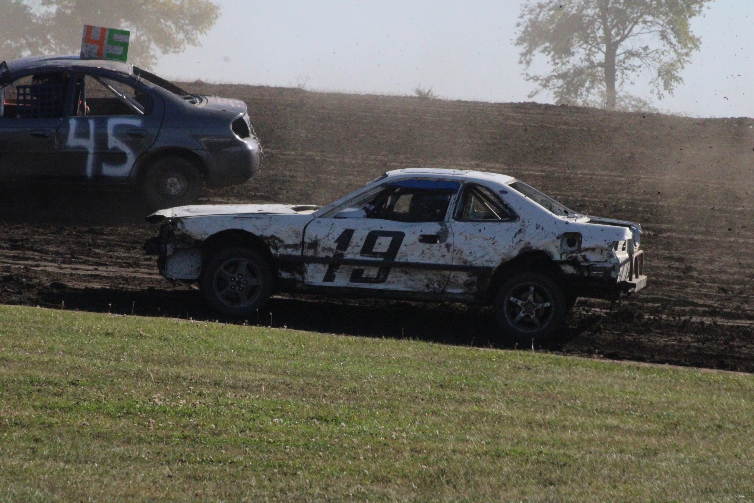 Damaged race car with the number 19, racing on a dirt track, with a black car in the background marked with 45.