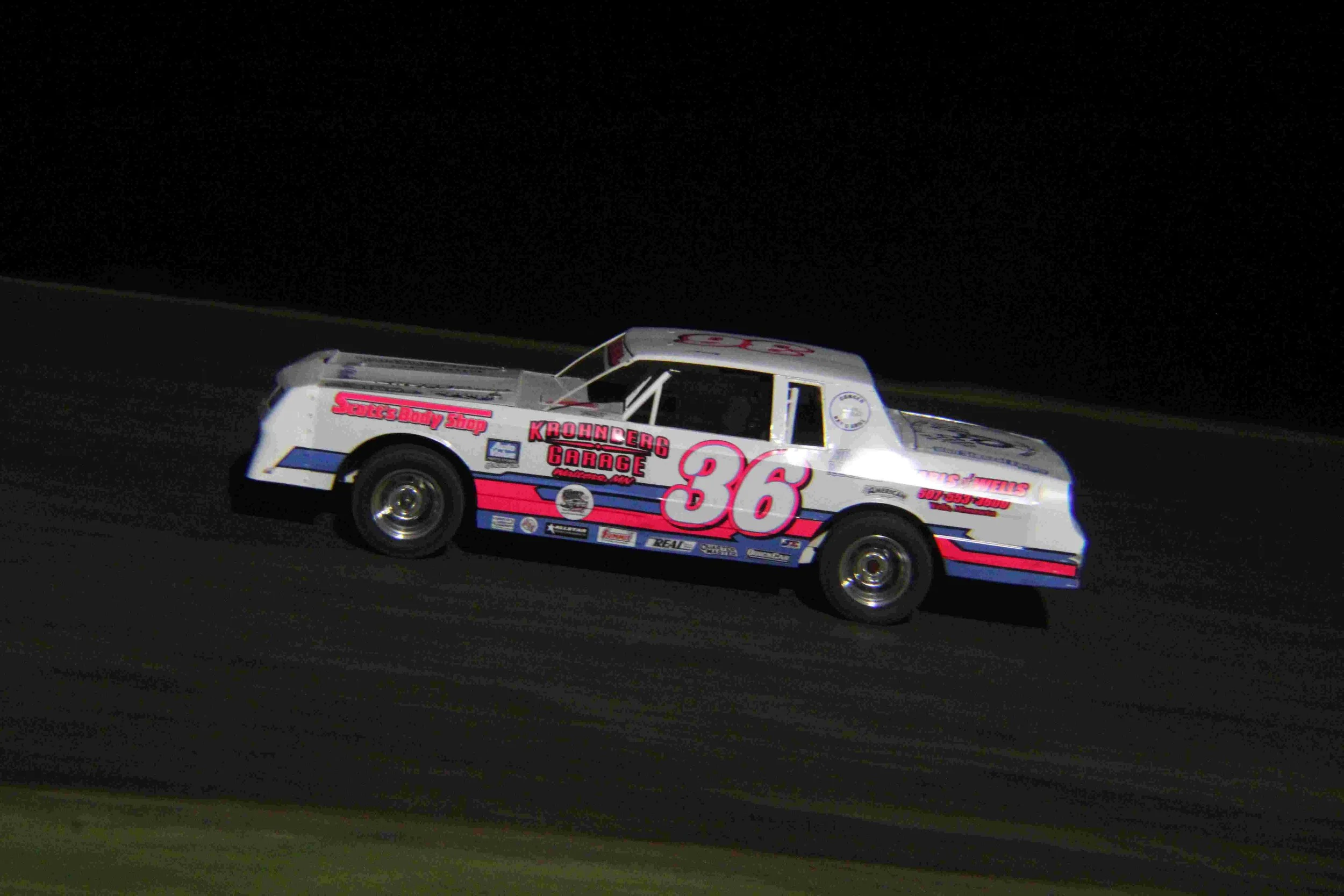 Nighttime image of race car number 36 on a racetrack, white with pink and blue accents, sponsored by Krohnberg Garage and Sports Body Shop.