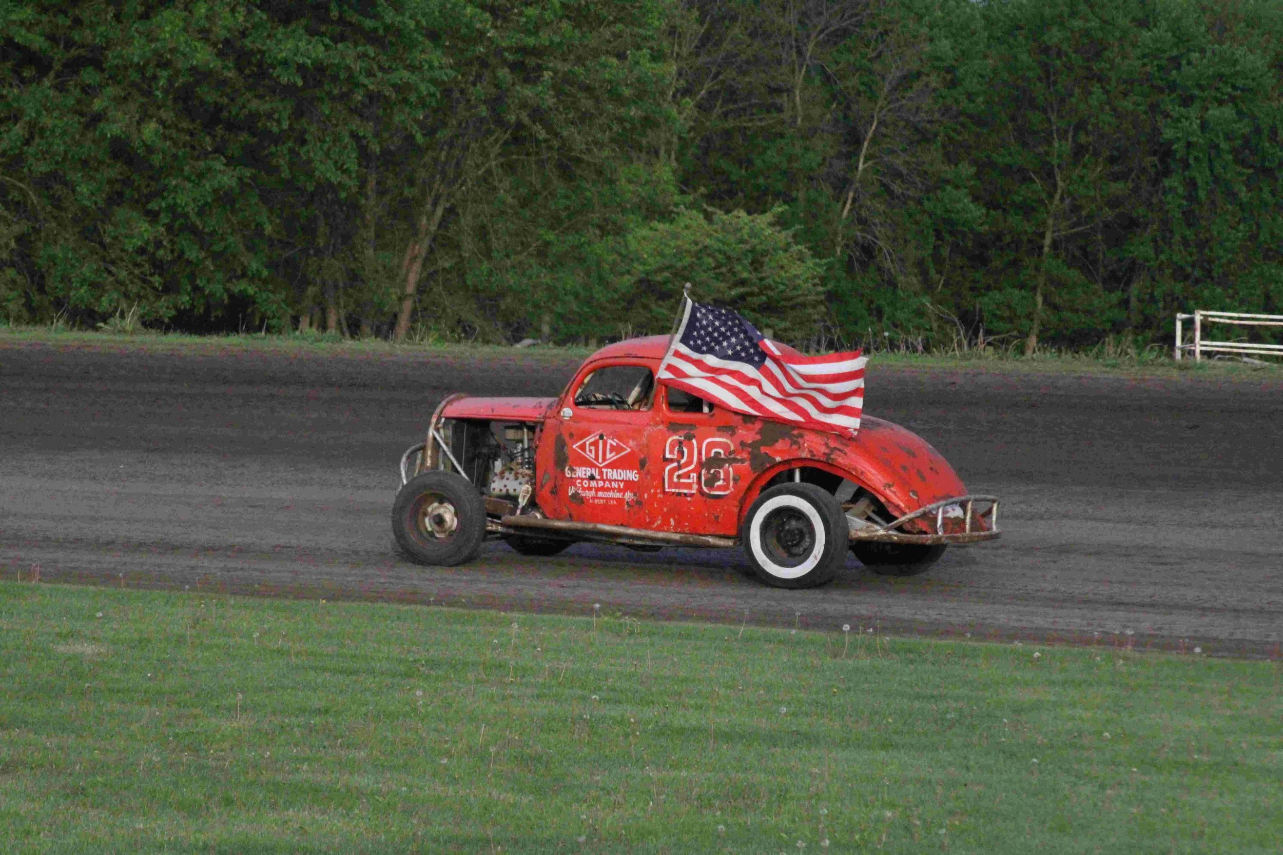 A vintage race car with the number 26 on the side, painted in red with visible rust spots, racing on a dirt track. An American flag is mounted on the car.