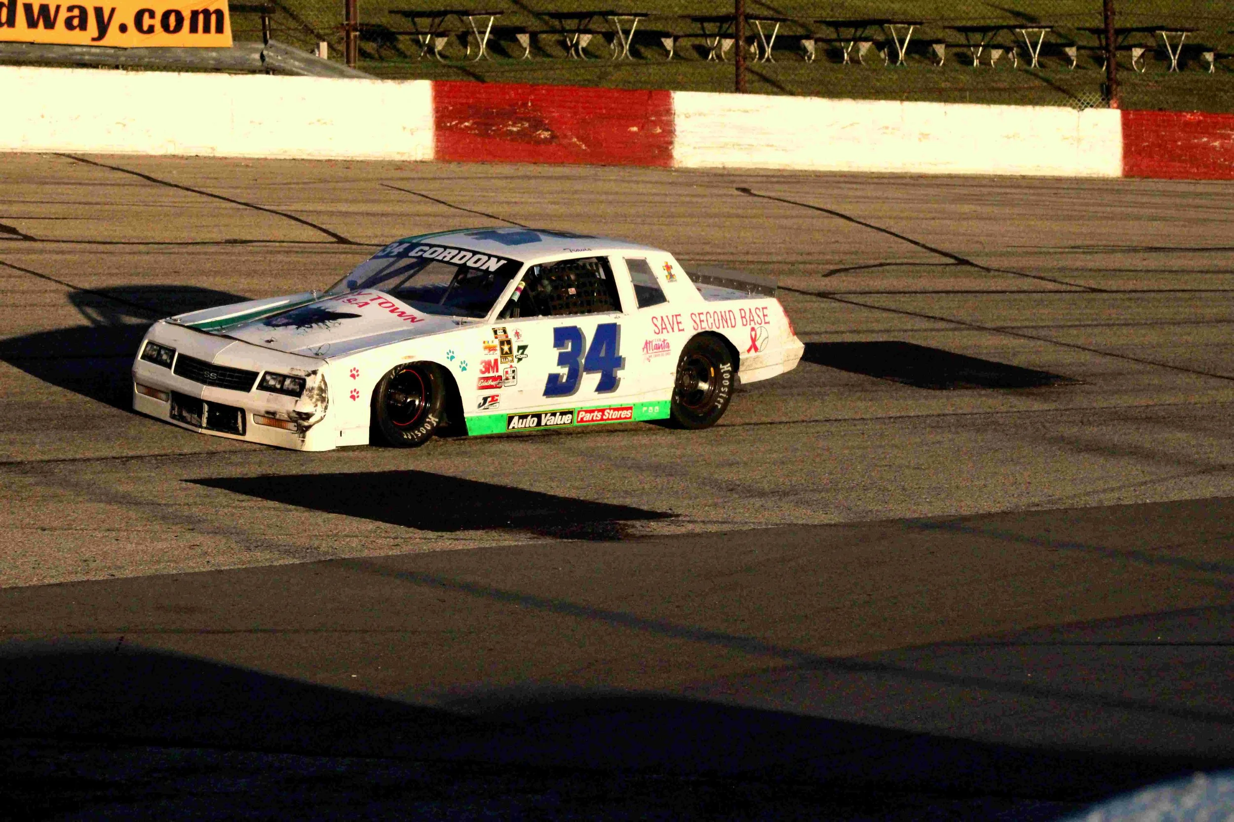 A white stock car with number 34 racing on a dirt track during a race, with red and green sponsor decals, and a red and white barrier in the background.