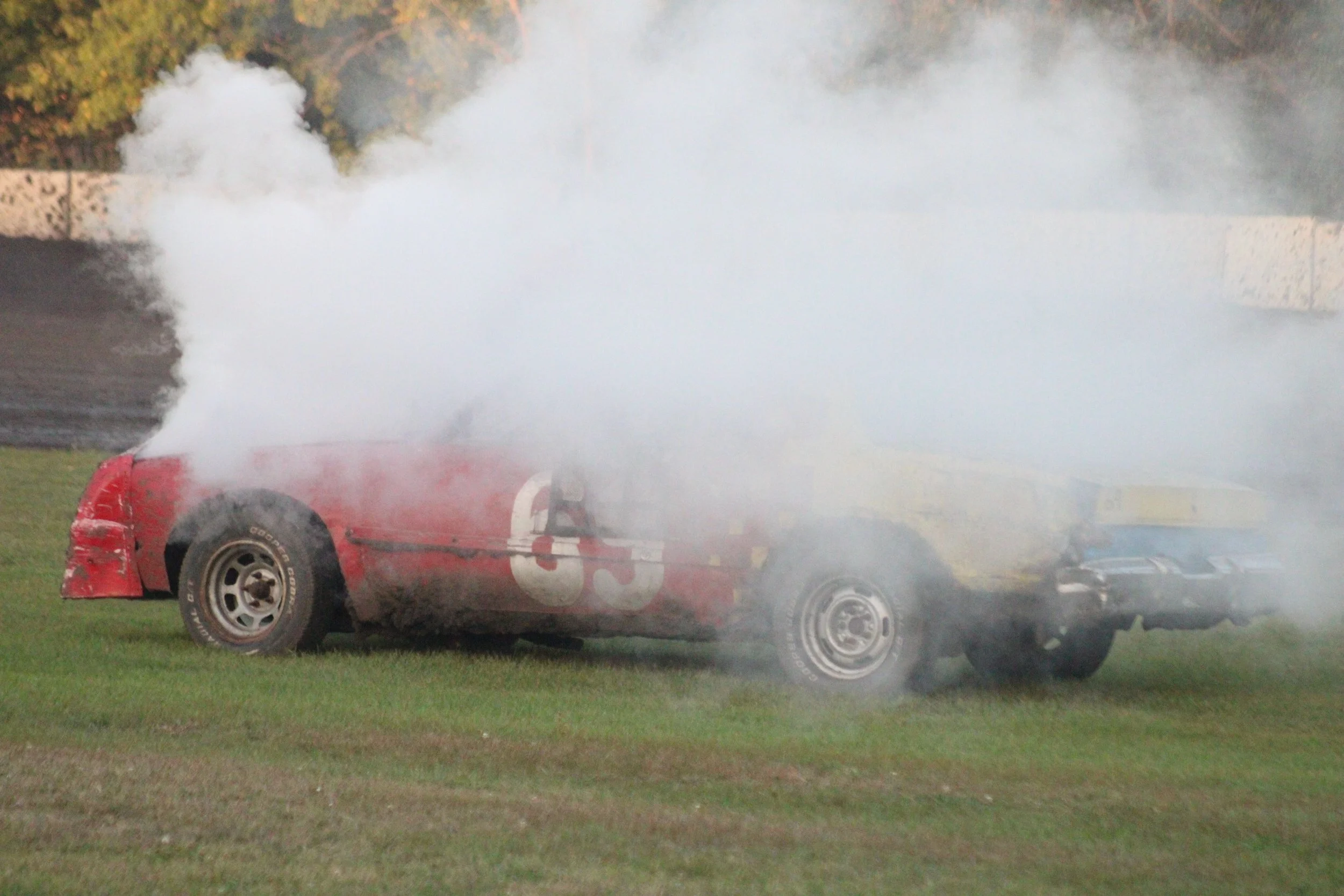 A red race car with the number 5 on the side is emitting white smoke in a grassy area.