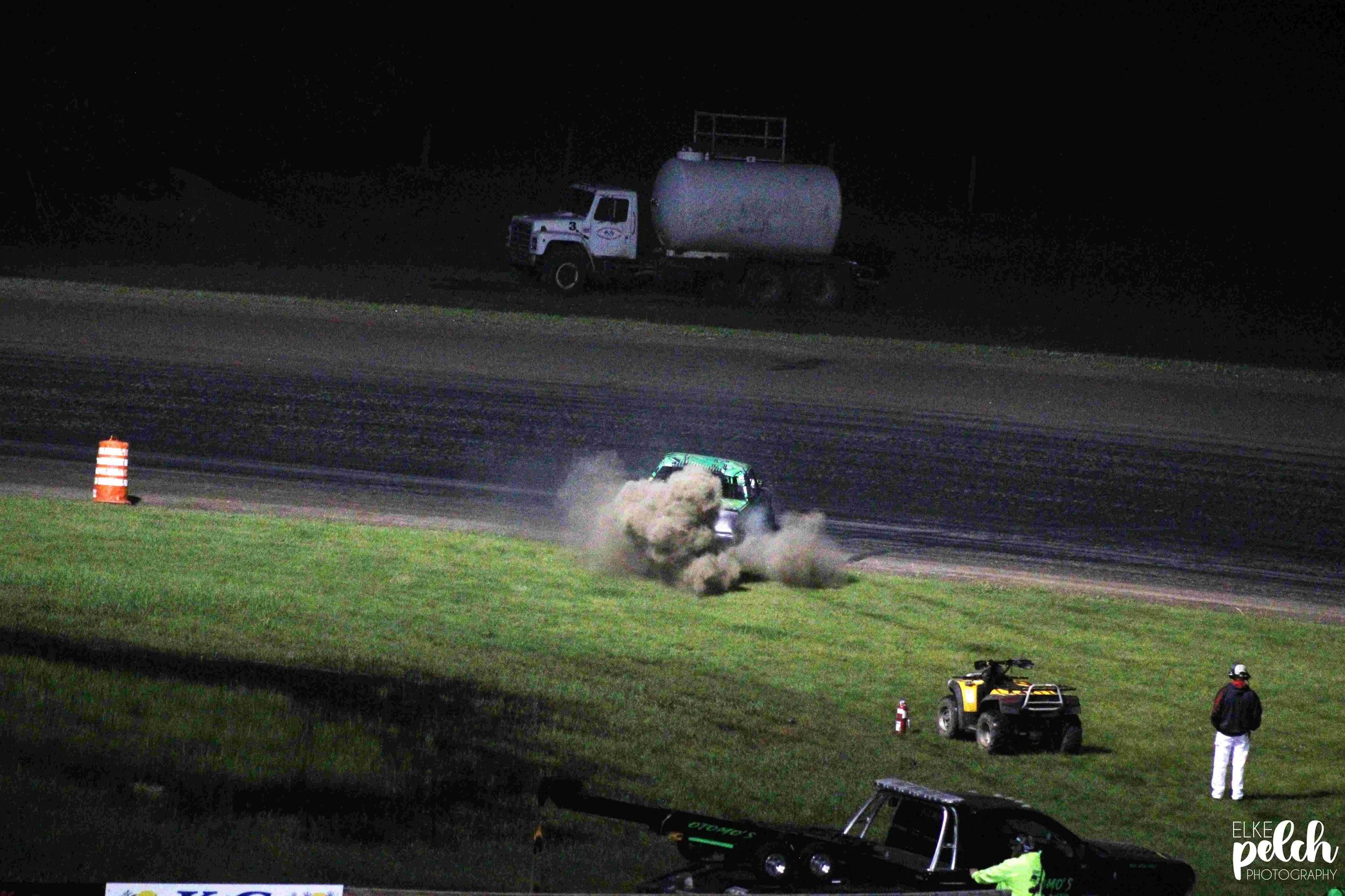 A nighttime scene at a racing event showing a green car crashing, kicking up dust and dirt on a dirt track. Two ATV vehicles and a person standing nearby are also visible on the grassy area. A large truck is parked on the darker background.