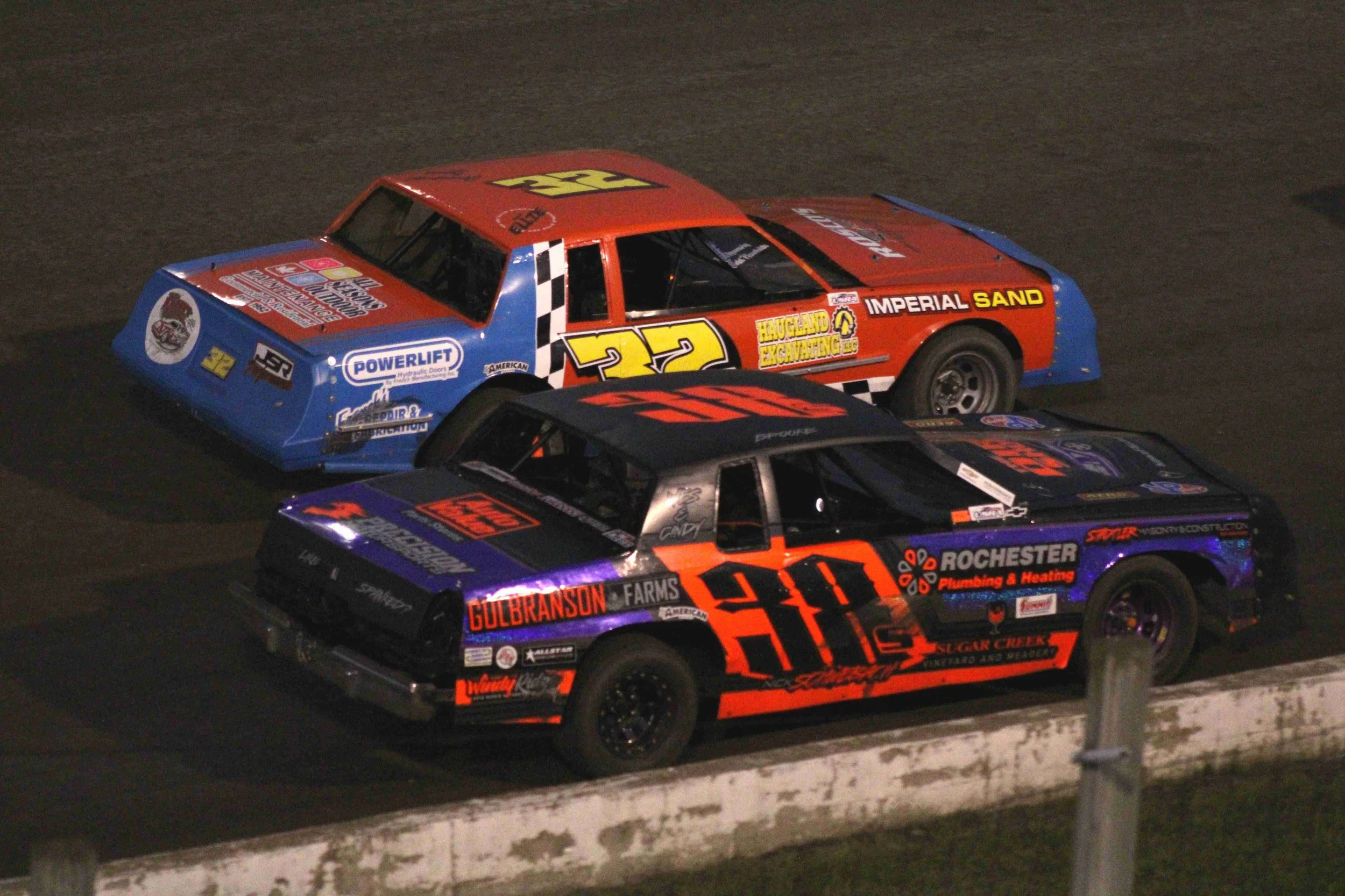 Two race cars on a dirt track, one with the number 22 in red and yellow on a blue and red car, and the other with the number 50 in orange and black on a purple and black car.