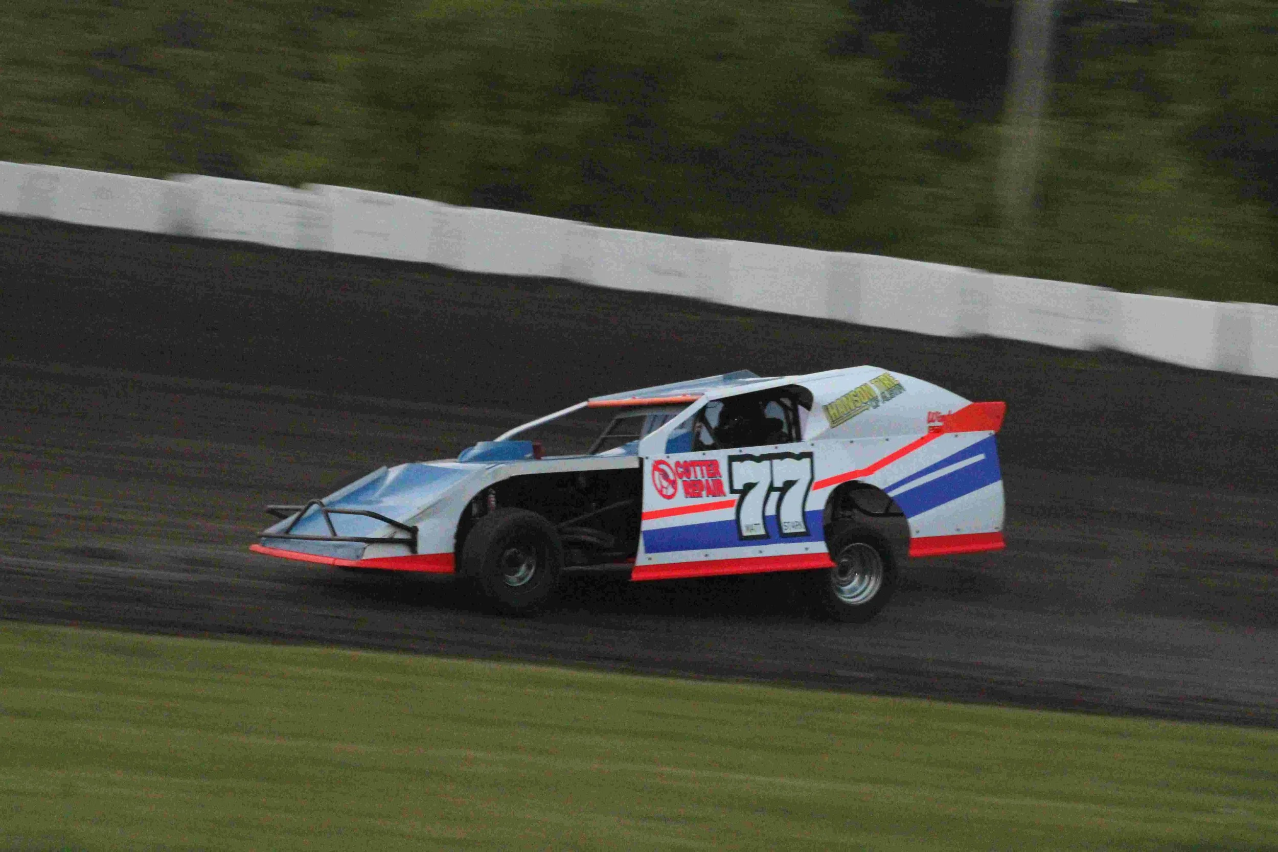 Race car number 77 speeding on a dirt track with a white barrier fence in the background.