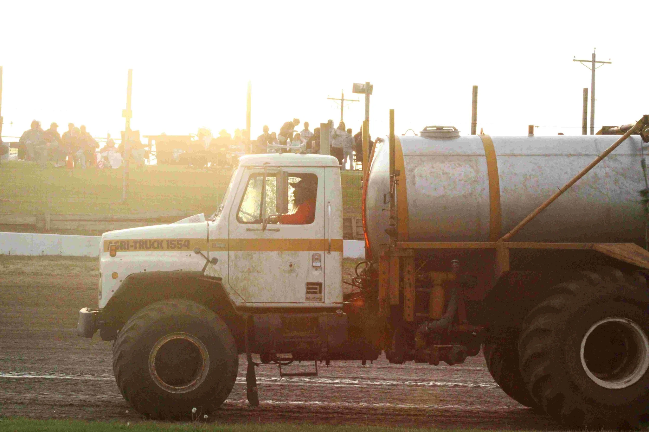 A large white industrial tank truck with yellow details on a dirt field during a sunset, with people sitting and standing on a grassy hill in the background.