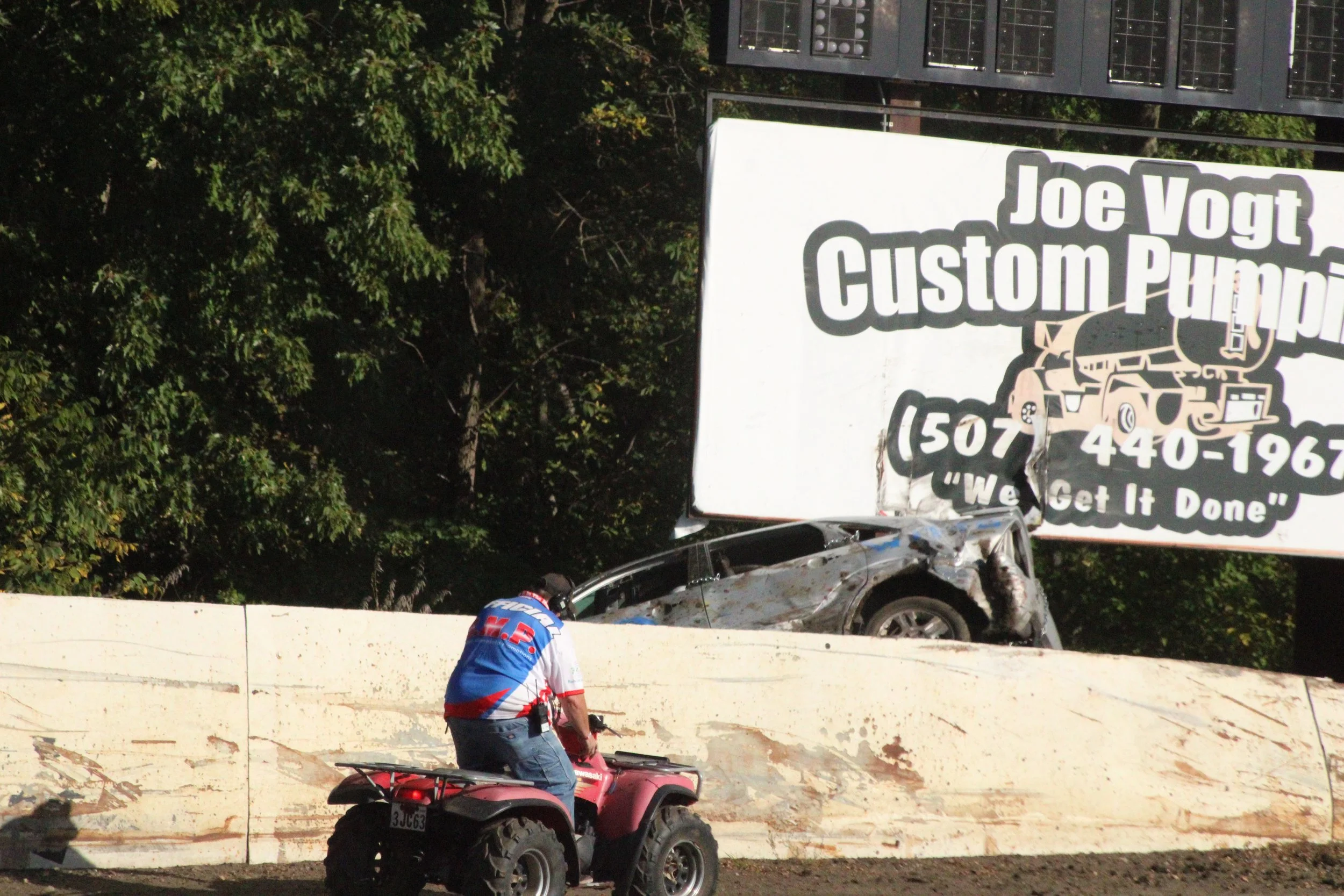A black vehicle crashed into a large billboard, with visible damage and debris around it, while a person on a red ATV rides past on the dirt track.