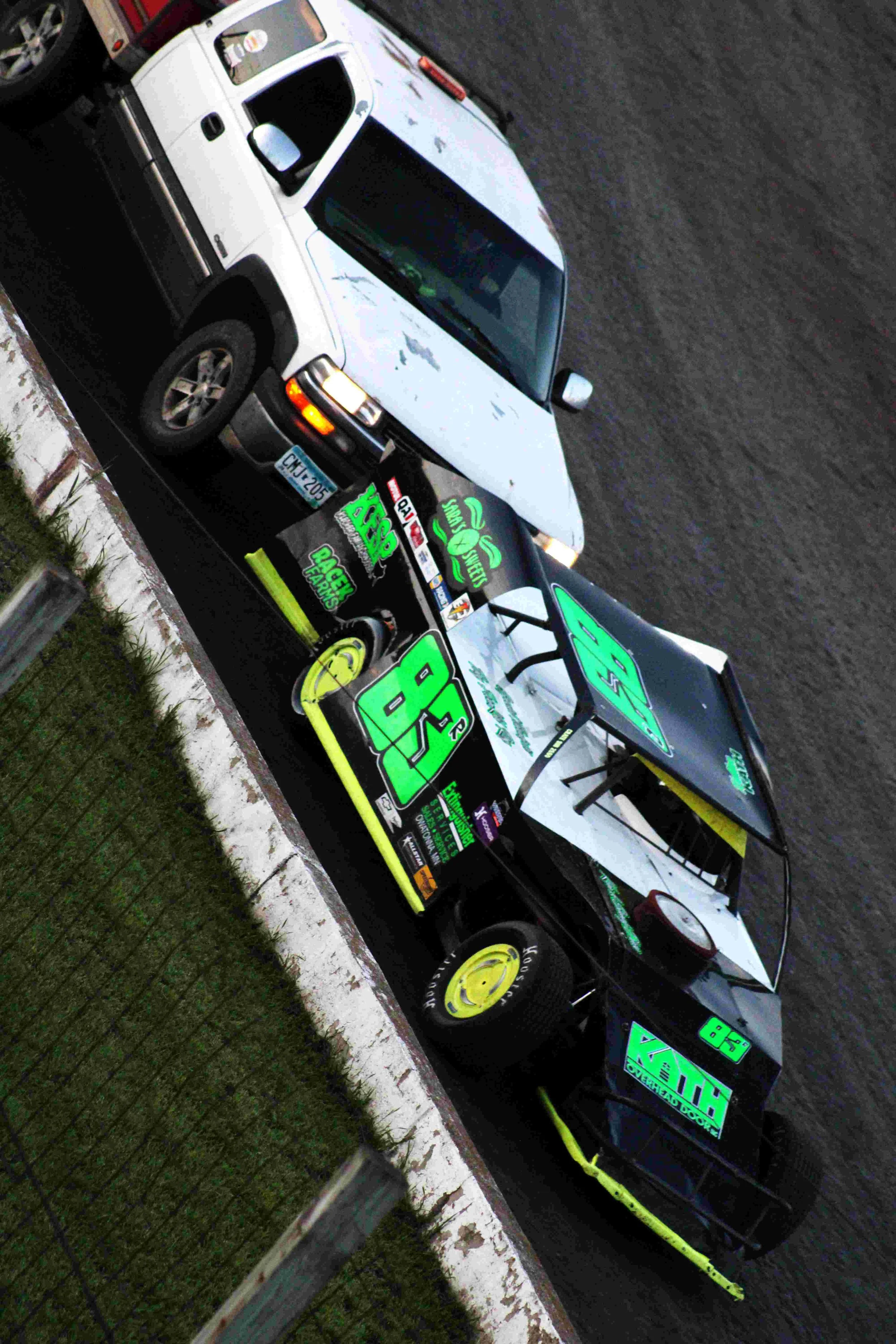 A black and white race car with the number 88 on its side, parked next to a white pickup truck. The race car features bright neon green and blue decals and a large rear spoiler.