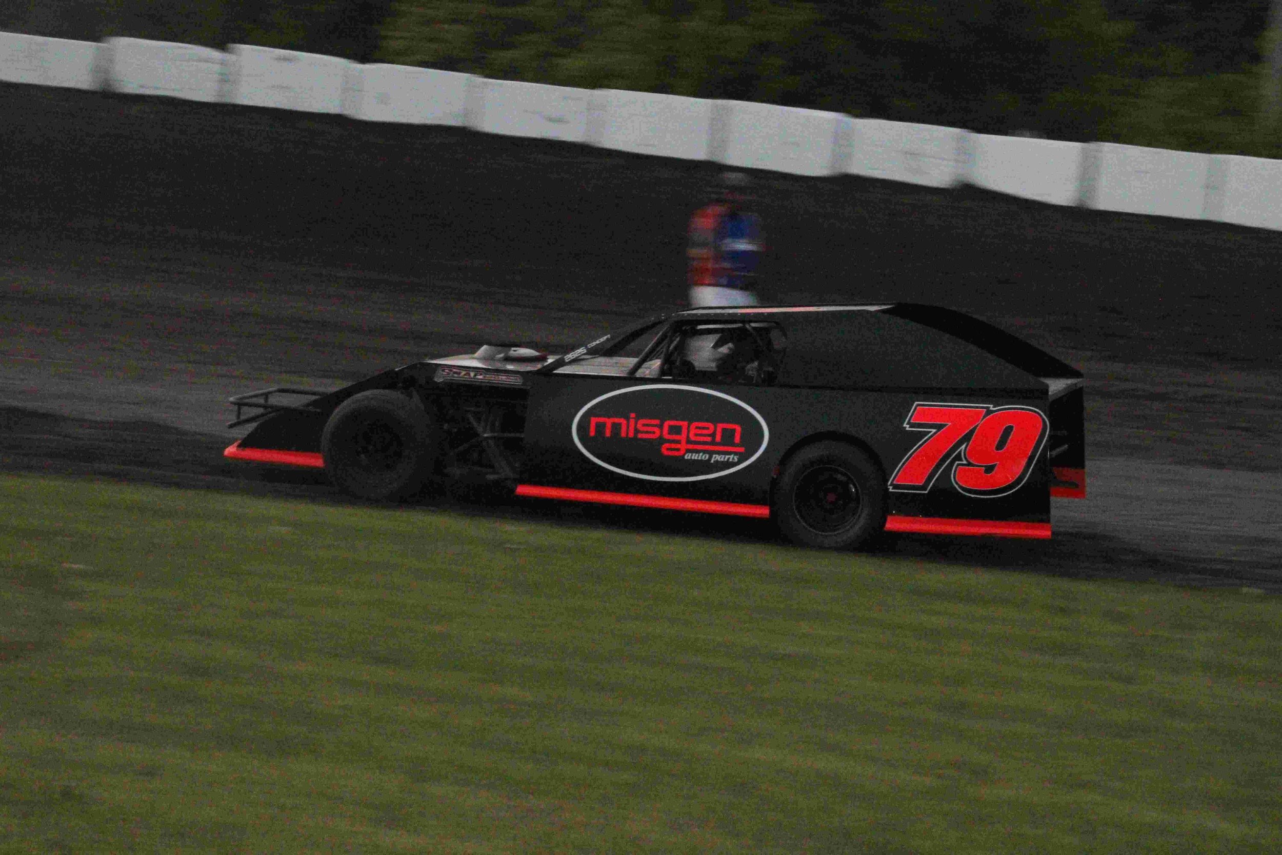 Race car driving on a dirt track, black with red accents, number 79, sponsored by Migen auto parts, with a person standing in the background.