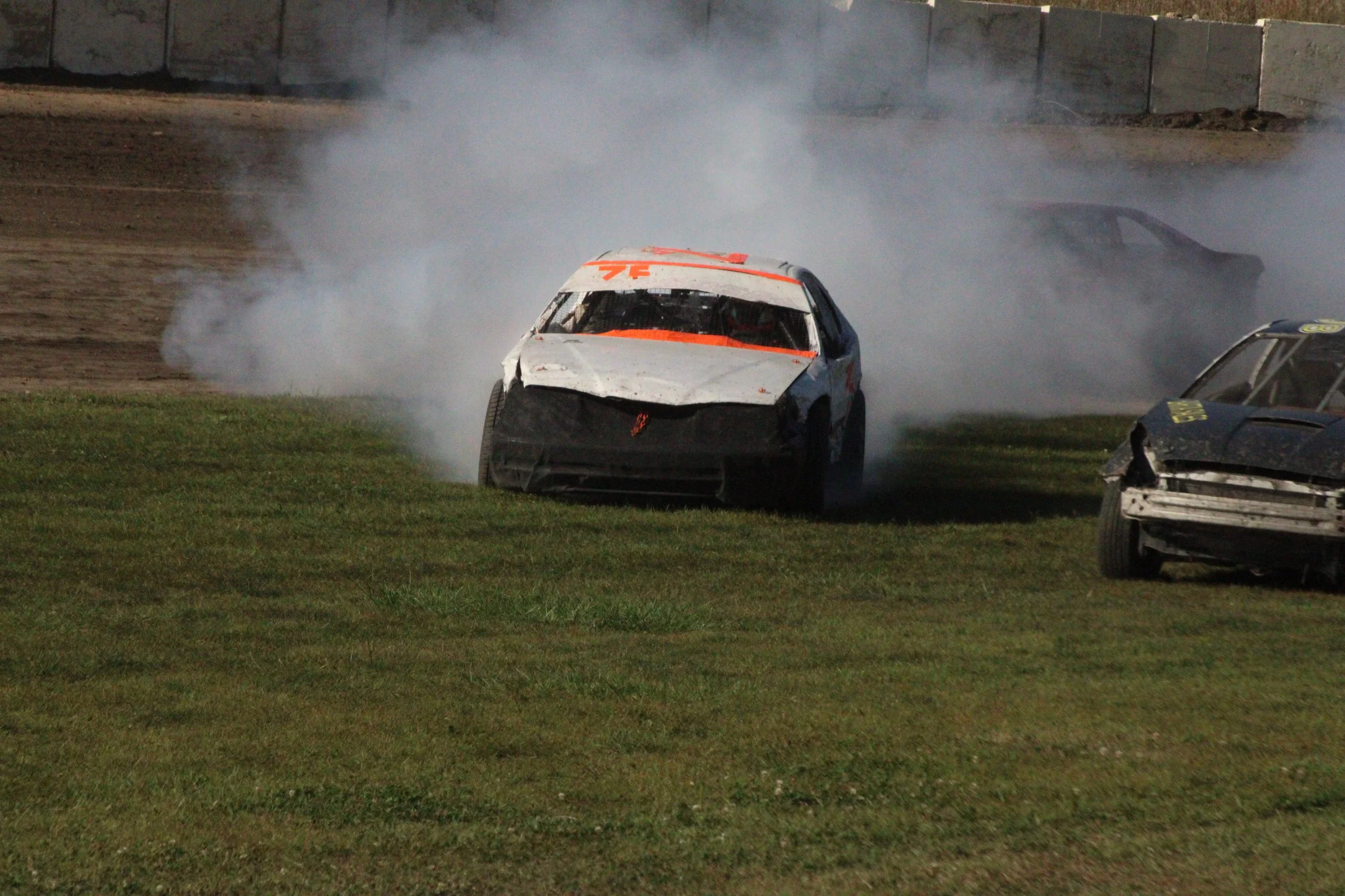 A race car crashing into the grass, producing smoke, during a dirt track race with other damaged cars nearby.