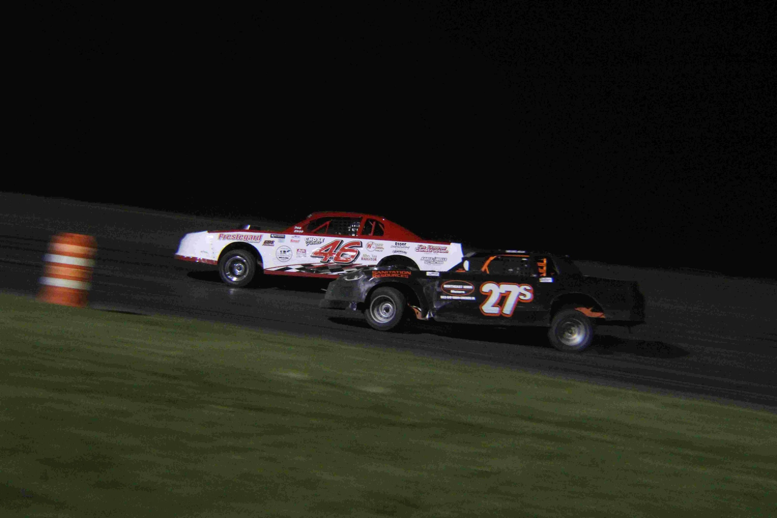 Nighttime dirt track race with two cars, one black with the number 27 and orange accents, and the other white with red details and the number 46, racing side by side.