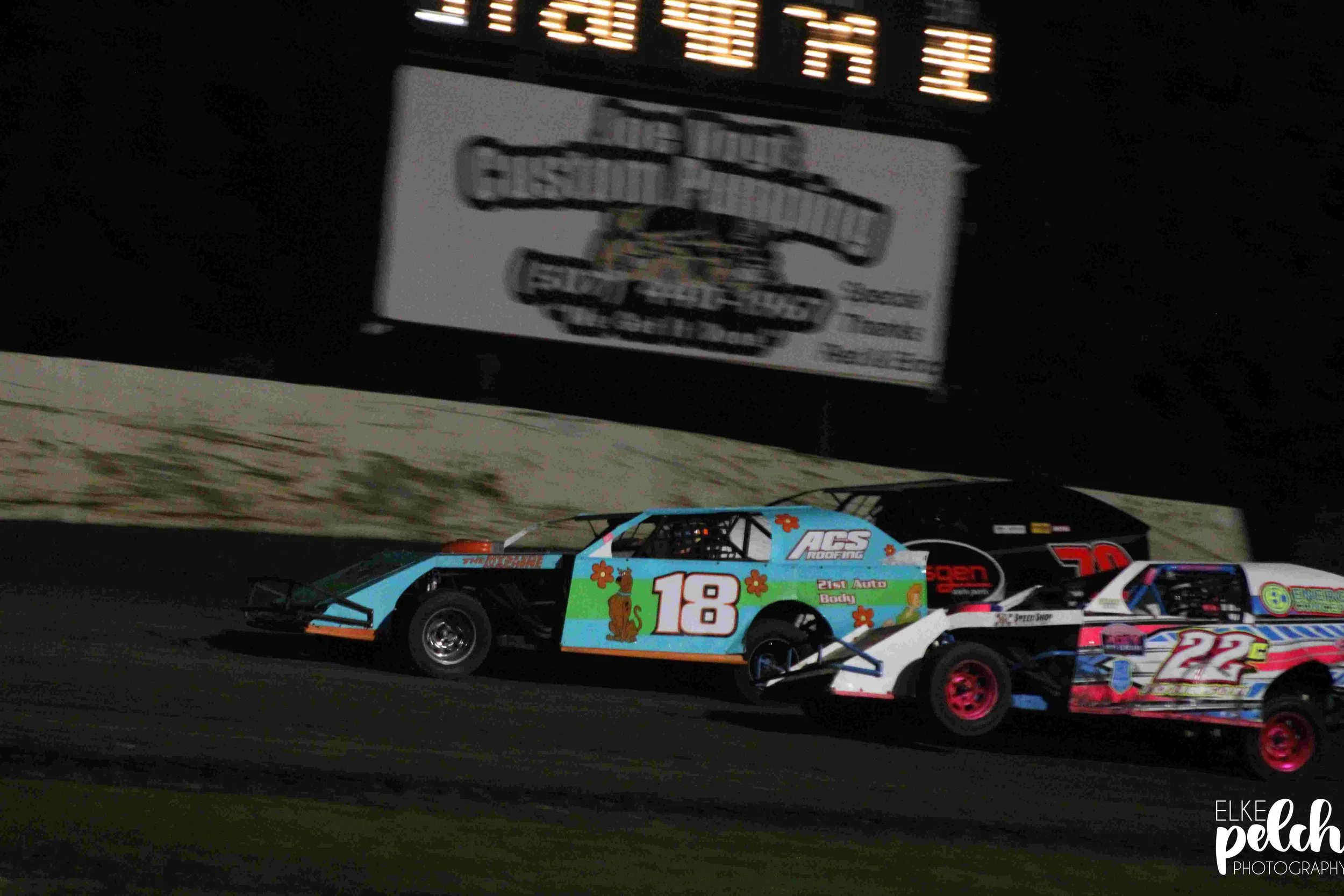 Three race cars competing on a dirt track at night. The cars are numbered 18, 22, and 70, with the 18 car featuring a colorful paint scheme with flowers and a dog illustration, the 22 car with blue, black, and pink accents, and the 70 car primarily b