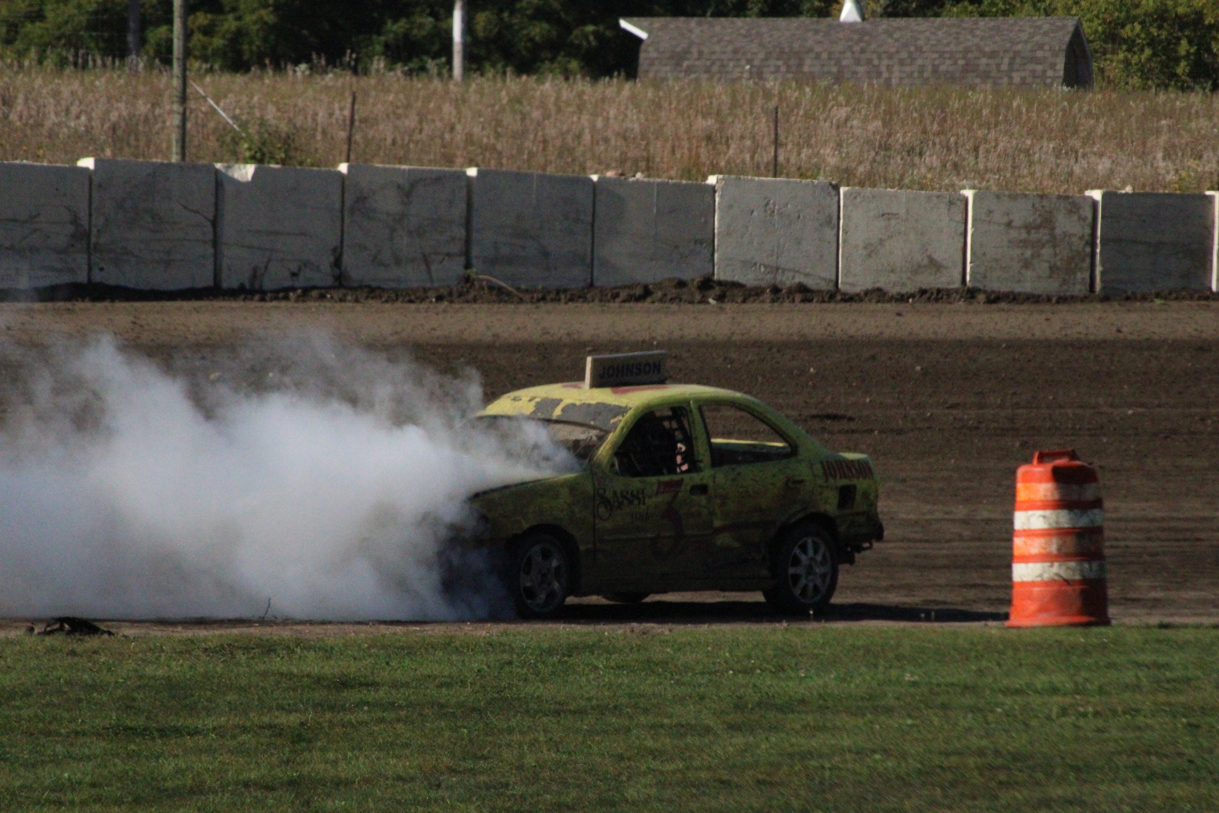 A green race car with the word 'Johnson' on the rear wing, emitting smoke, on a dirt track with a large orange traffic barrel nearby, and a concrete barrier in the background.