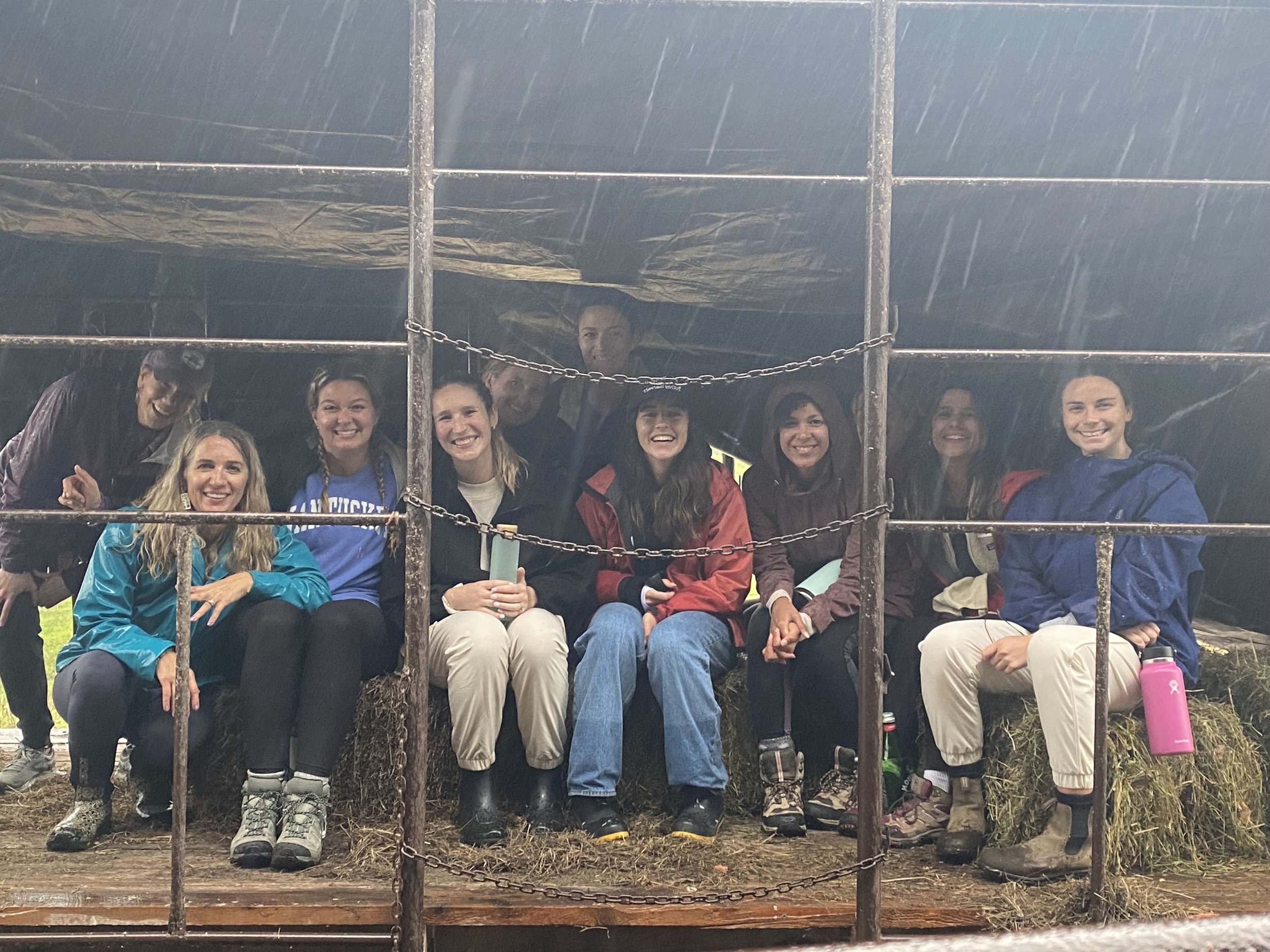 Group of people sitting and smiling on hay bales inside a sheltered area during rainy weather.