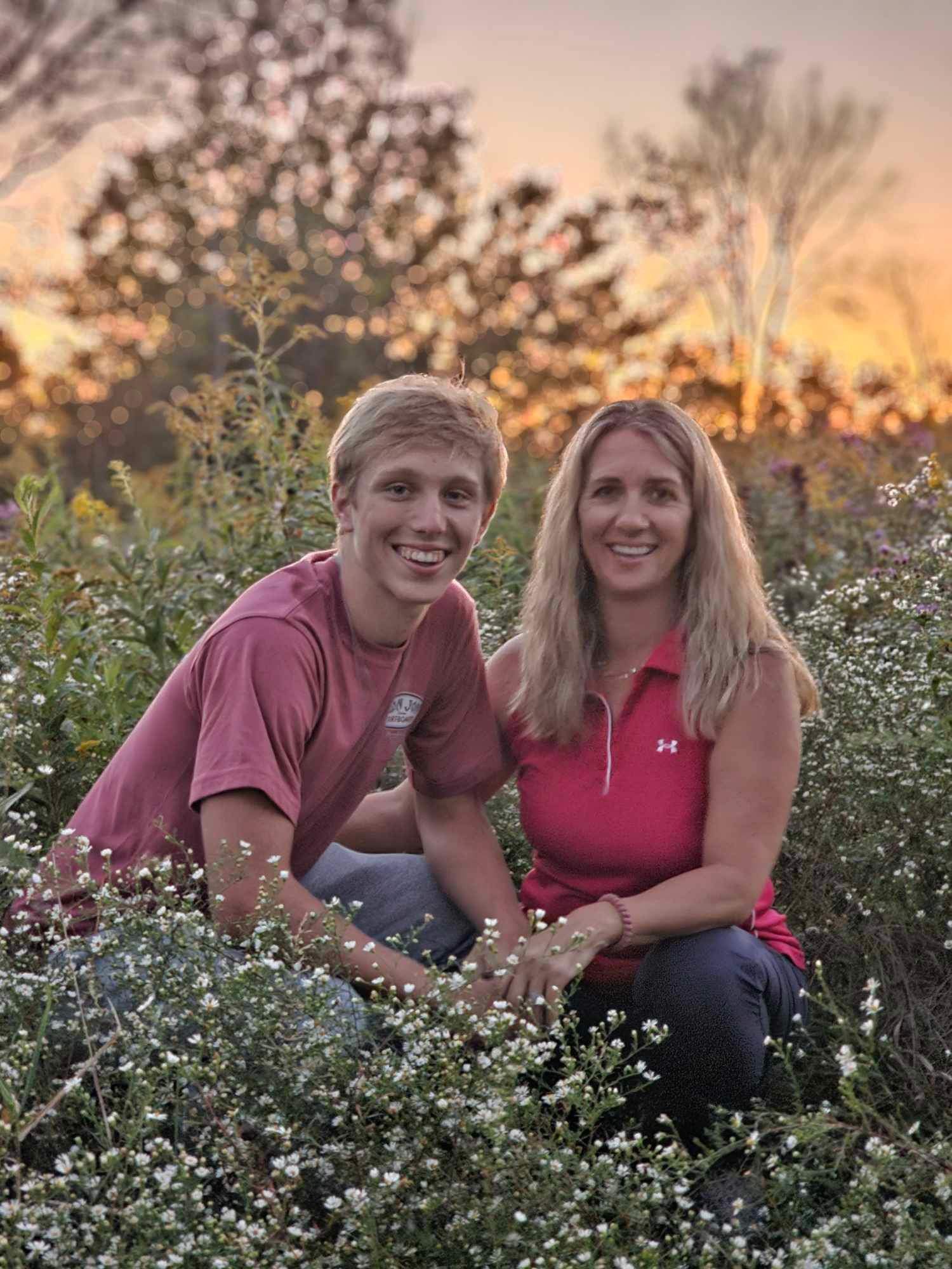 A smiling young man and a woman sit among white flowers in a garden during sunset, with trees and a colorful sky in the background.
