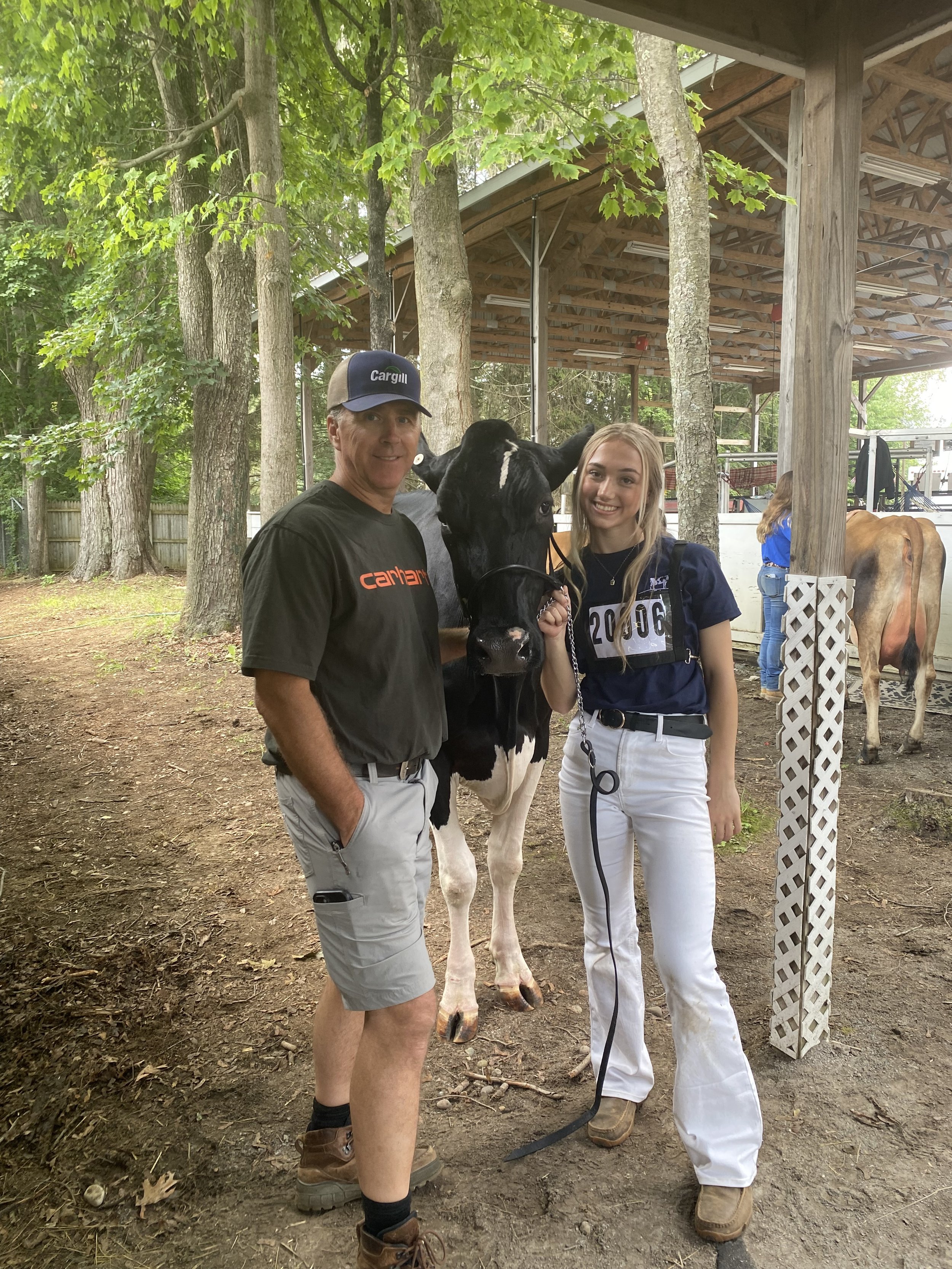 A man and a woman smiling next to a young calf with a person in the background, outdoors with trees and wooden structures.