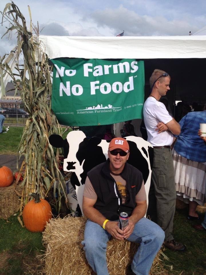 A man wearing a CAT baseball cap and sunglasses sitting on a hay bale in front of a cow print backdrop at a farm event. There are pumpkins and dried corn stalks on the ground. A green banner reads 'No Farms No Food' and a woman is partially visible o