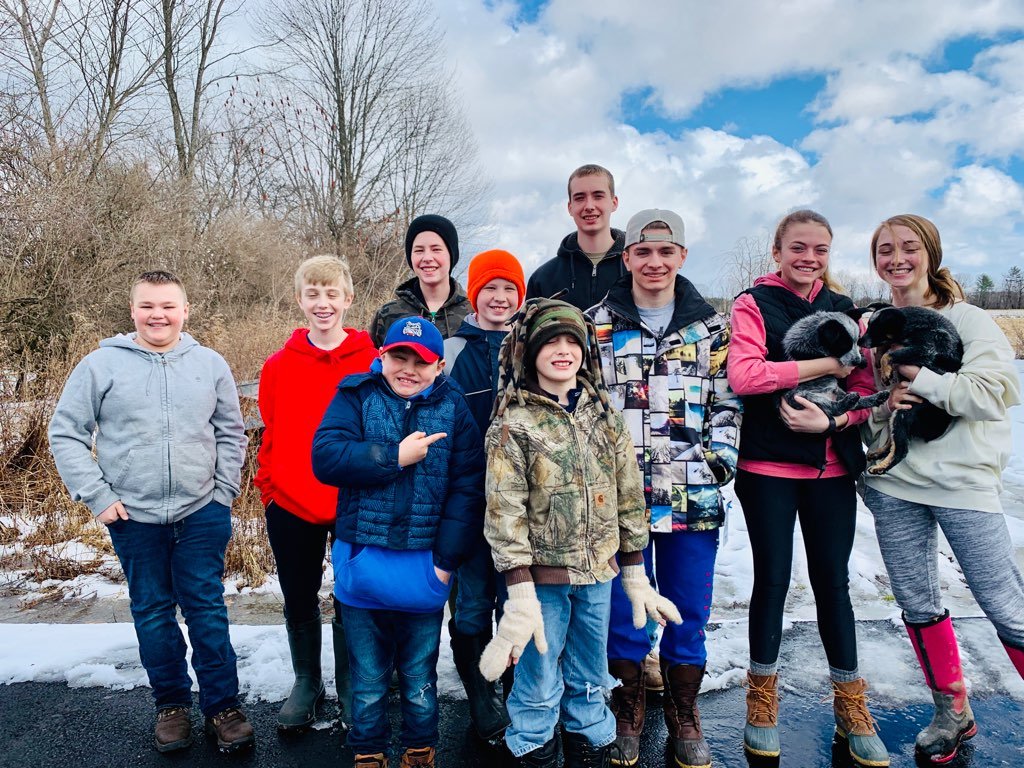 Group of children and teenagers outdoors in winter, some holding puppies, with a snowy landscape, leafless trees, and a partly cloudy sky in the background.