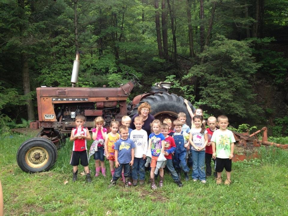 Group of children and a woman standing in front of a rusty tractor in a wooded area.