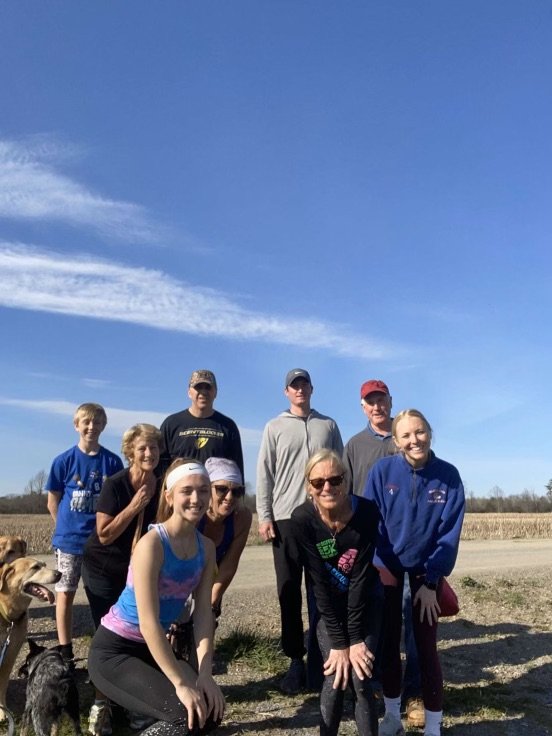 A group of nine people, including children and adults, standing outdoors on a sunny day with a clear blue sky and some clouds, in an open field with a dog in the foreground.