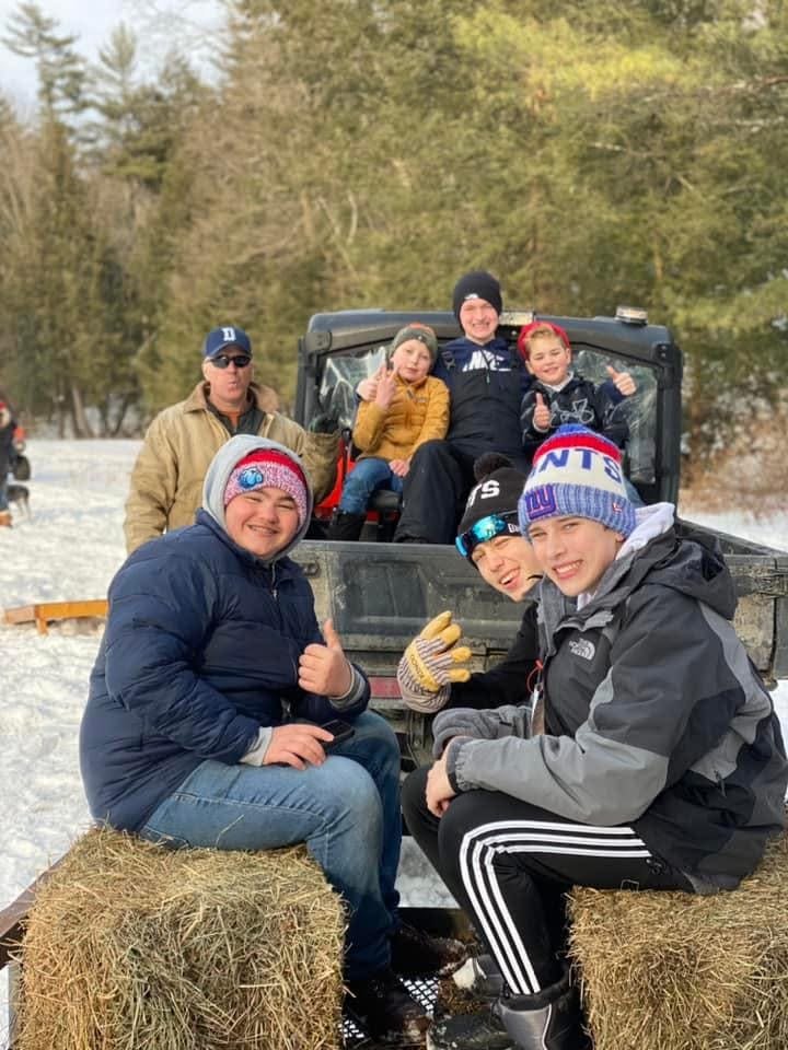 Group of children and an adult in a snowy outdoor setting, sitting on hay bales and a vehicle with a forest background.