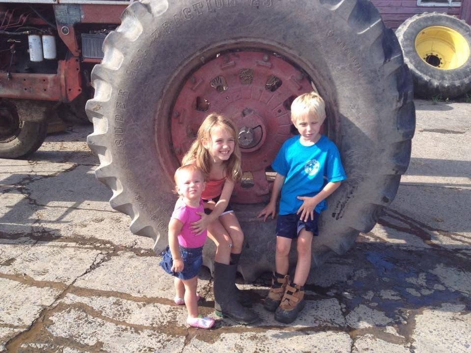 Three children posing in front of a large tractor tire outdoors, with two of them inside the tire and one standing in front of it.