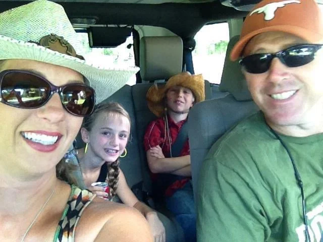 Family of four sitting inside a vehicle, dressed casually and wearing hats and sunglasses, smiling for a selfie.
