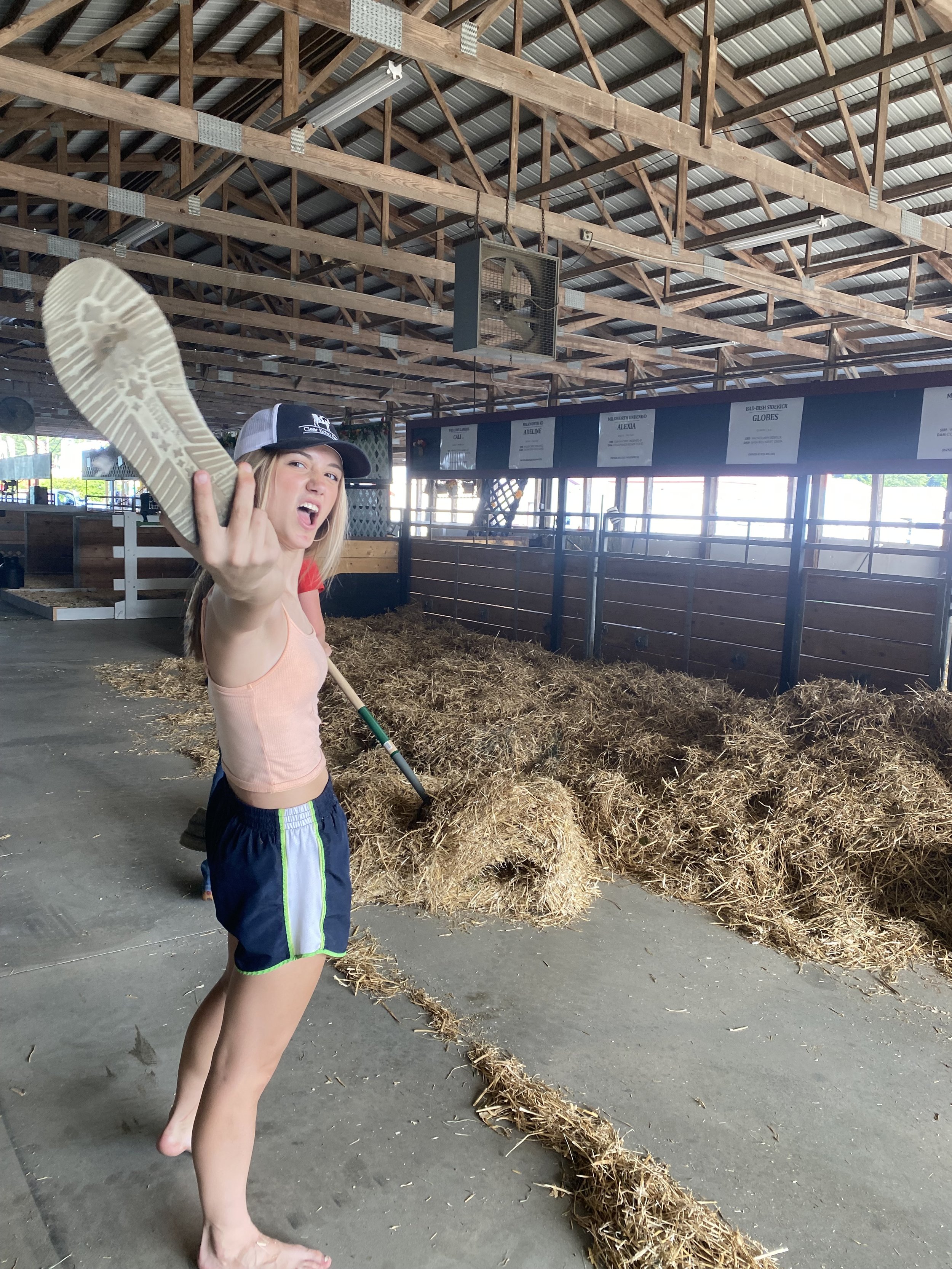 A woman barefoot, wearing a tank top, shorts, and a cap, is inside a barn or stable holding a shoe toward the camera with an aggressive or playful expression.