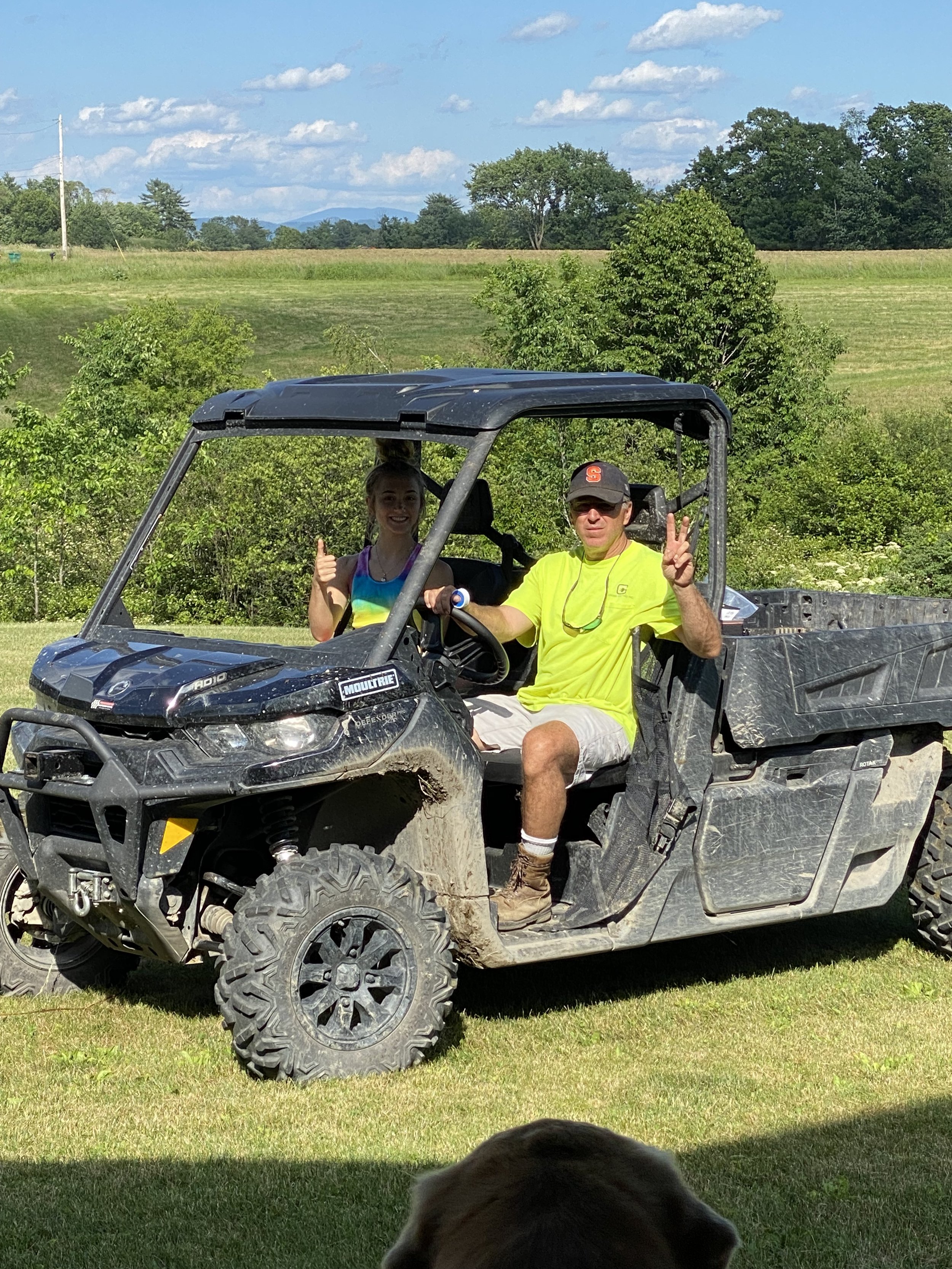 A man and a young girl sitting in a black utility vehicle outdoors on a sunny day. The man is wearing sunglasses and a gray cap, making a peace sign with one hand and giving a thumbs up with the other. The girl is smiling and giving a thumbs up. The