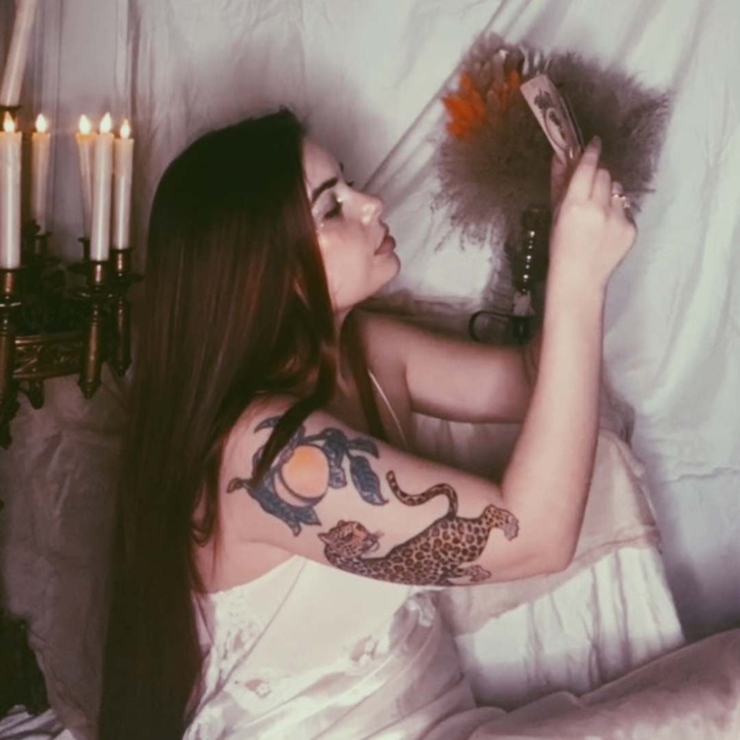 A woman with long dark hair, tattoos of a tiger, snake, and lion on her arm, lies on a bed taking a selfie with her phone. Candles are lit in the background on a wall sconce.